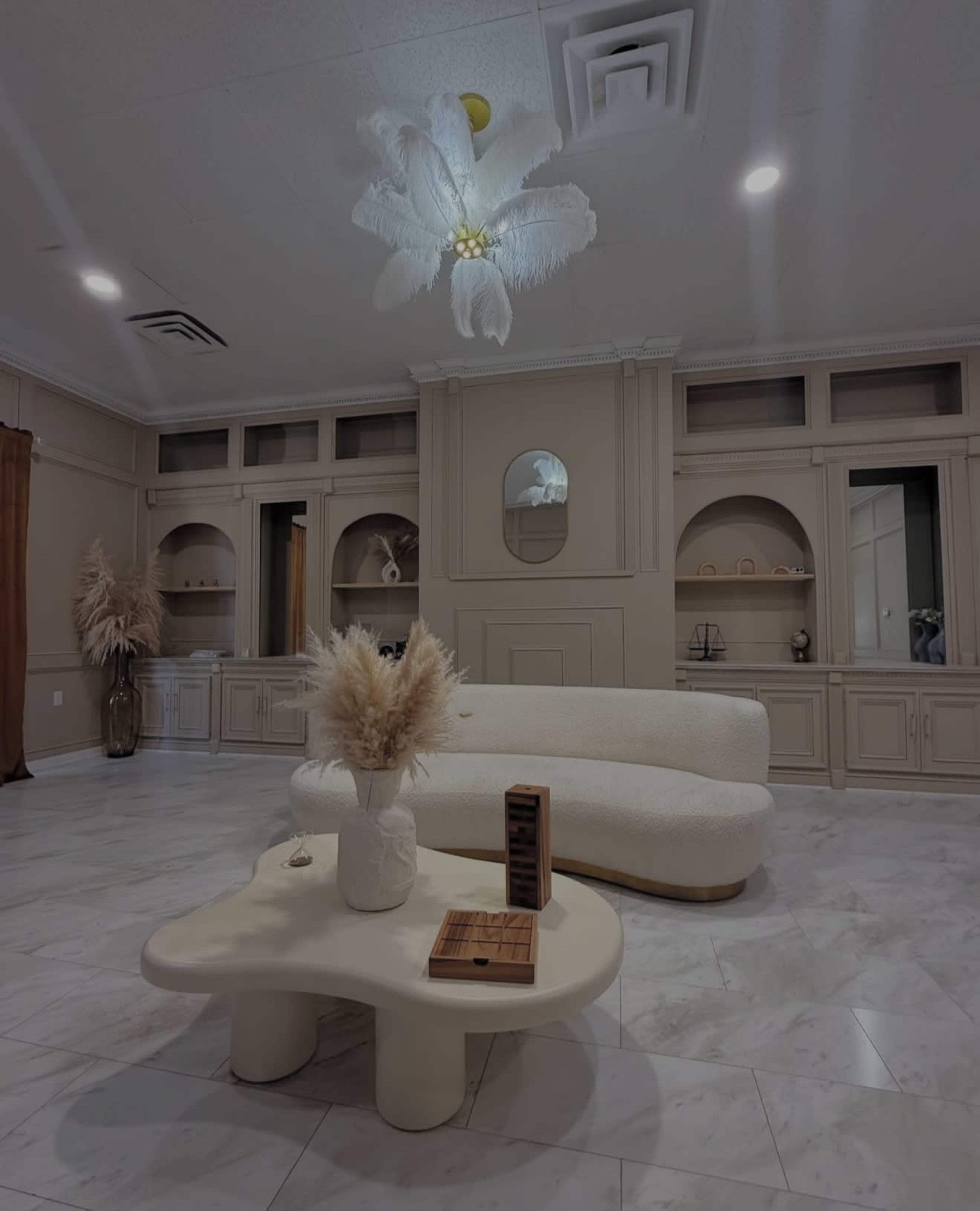 The image shows a modern, neutral-toned living room with a plush curved sofa, a sculptural coffee table, and decorative elements including dried plants and minimalistic decor on the shelves.