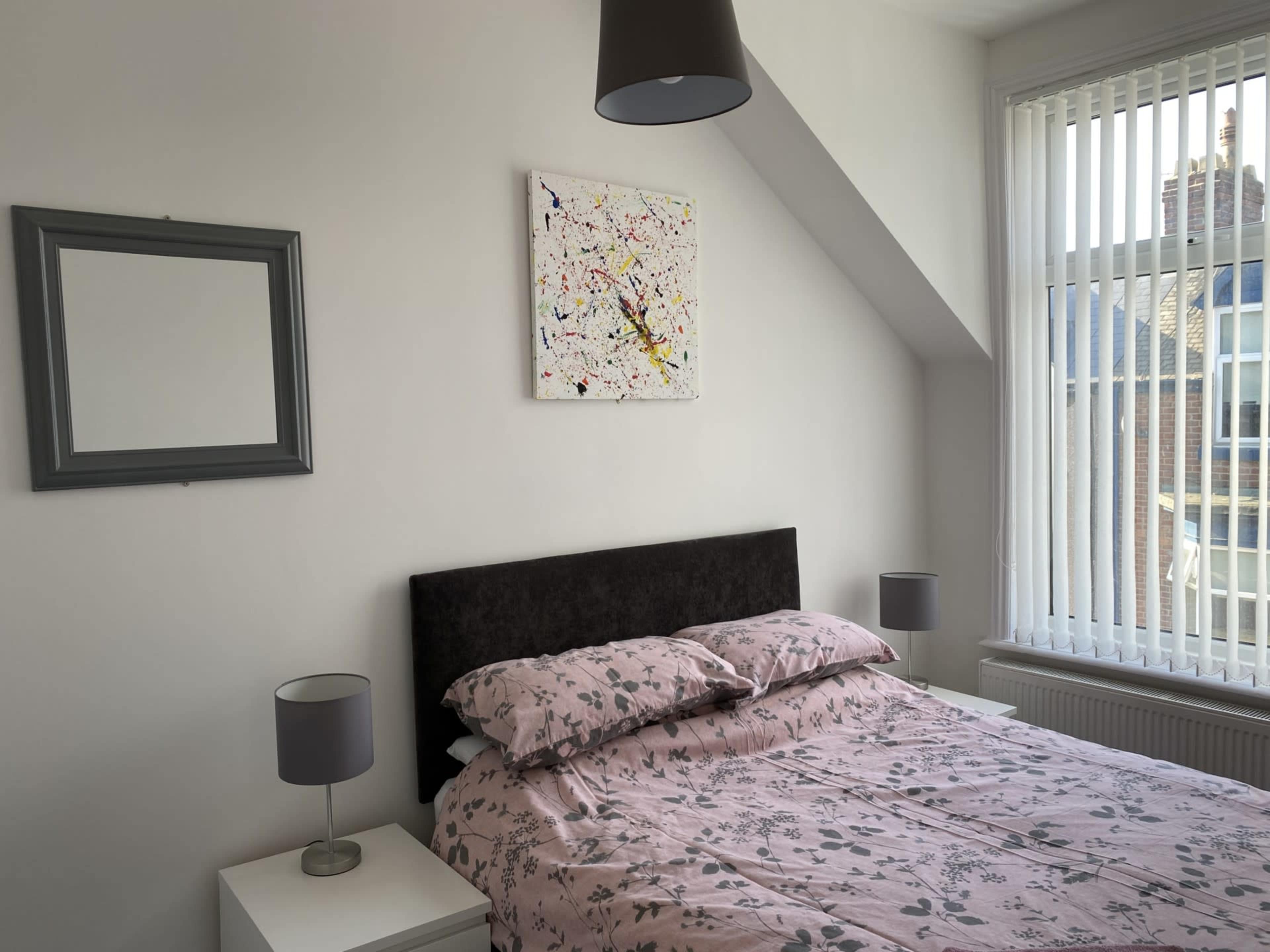 A bedroom featuring a bed with a floral-patterned duvet, a wall-mounted painting, and a bedside table with lamps.