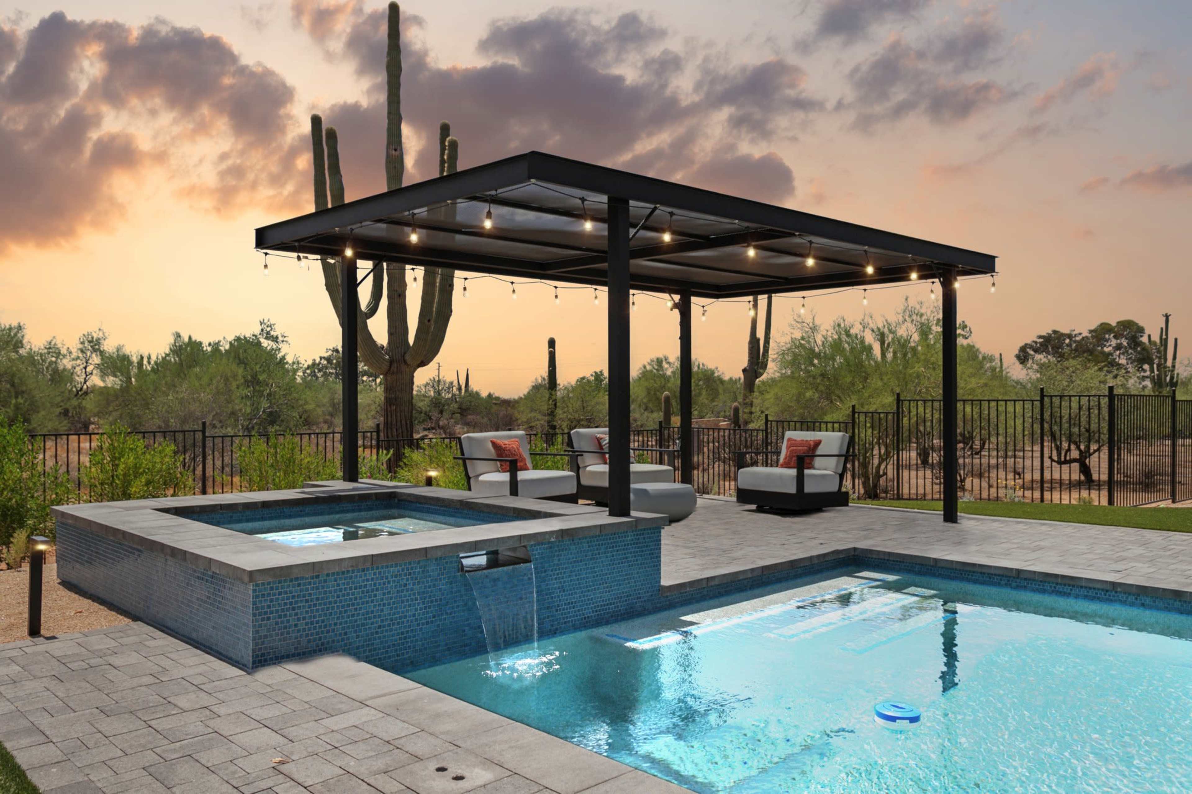 The image shows a modern backyard with a swimming pool, a hot tub, and a covered seating area, set against a sunset and surrounded by desert vegetation.