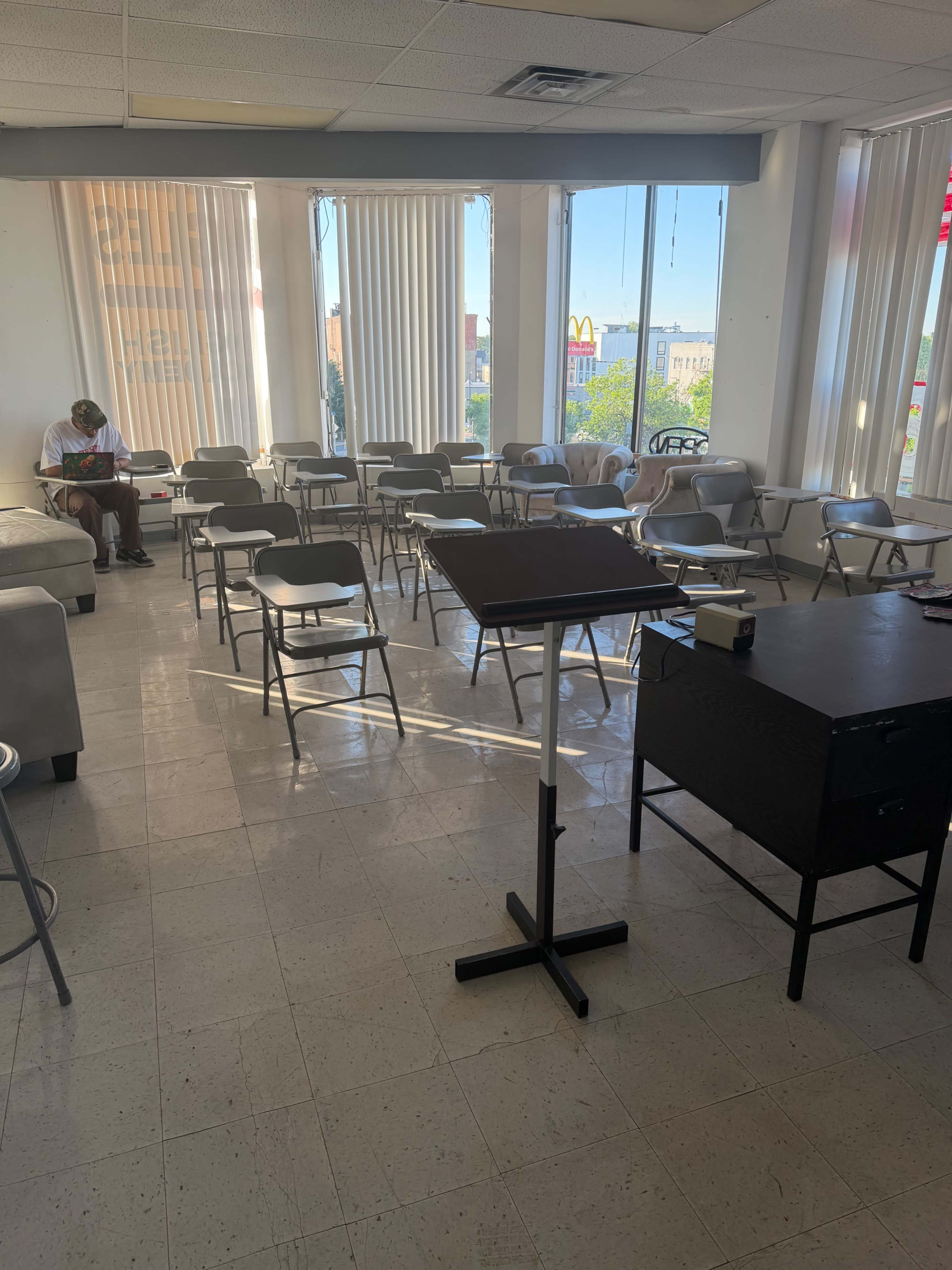 The image shows a sparsely populated classroom with rows of chairs and desks, and a person sitting at the back of the room.