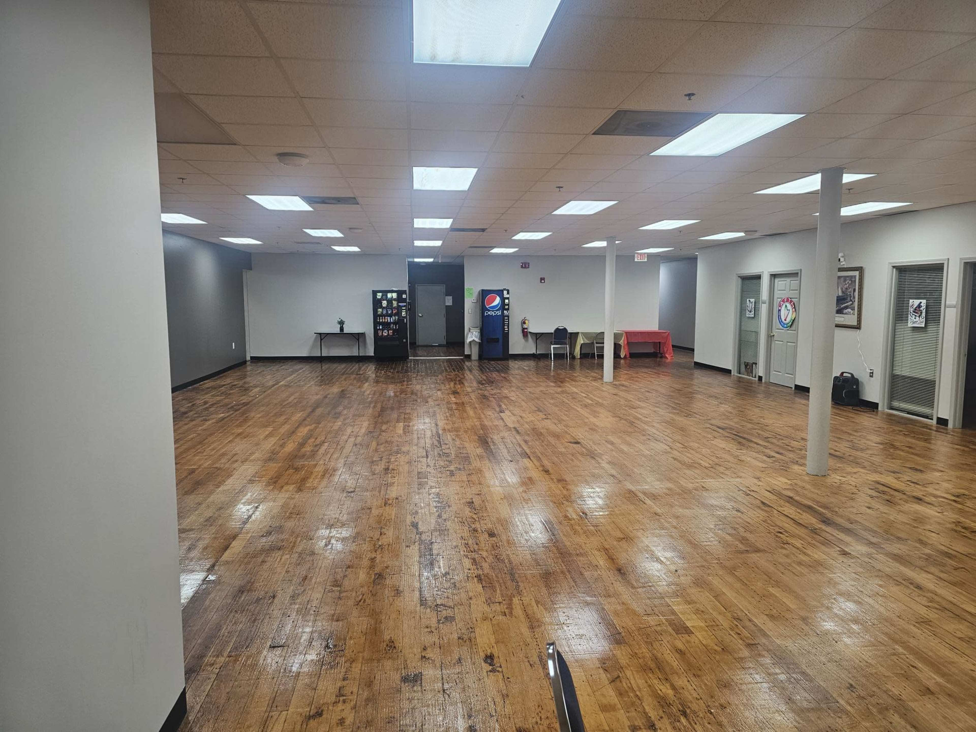 The image shows a spacious, empty room with wooden flooring, overhead lights, and a few pieces of furniture against the walls.