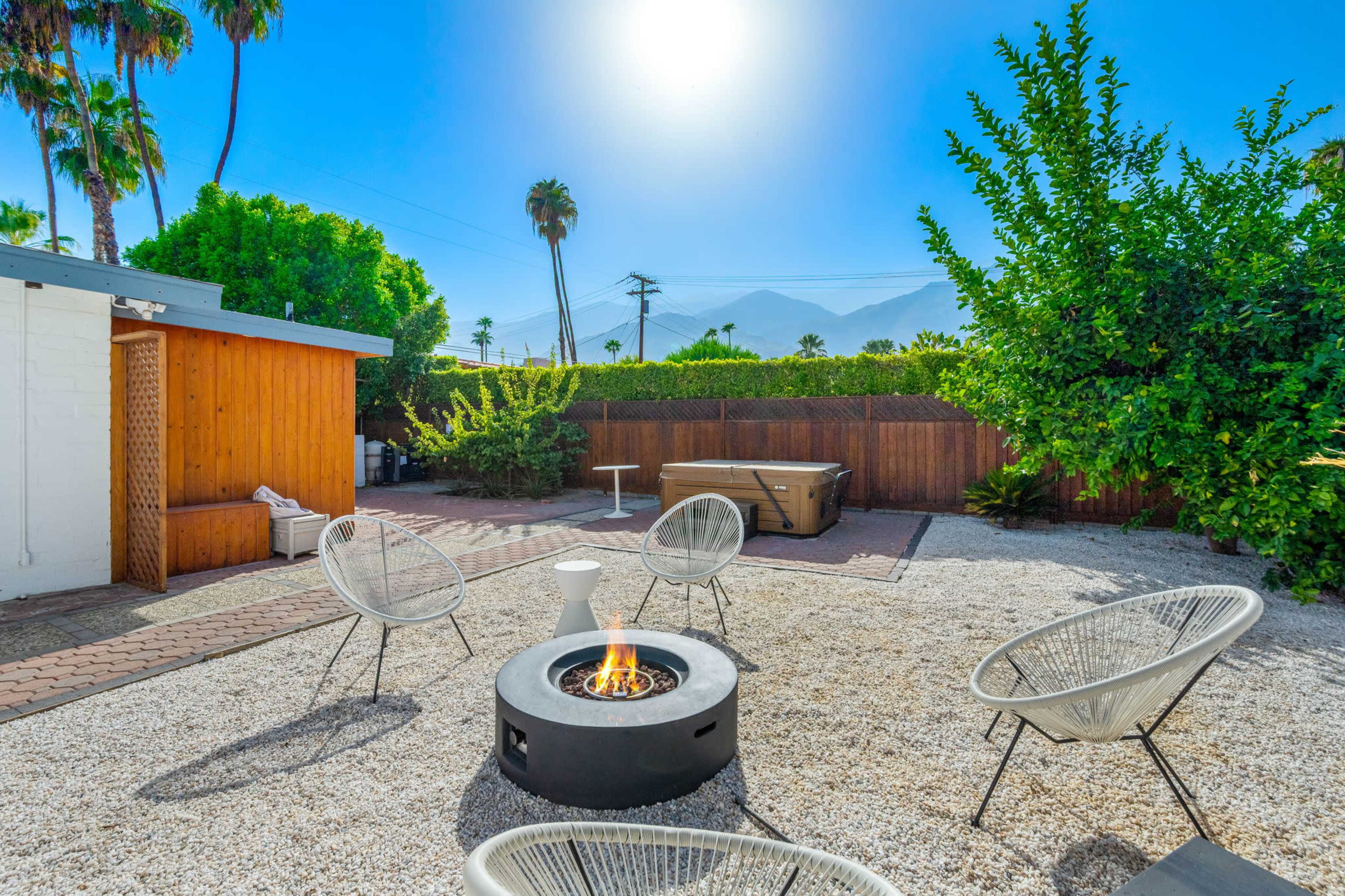 A circular fire pit surrounded by four chairs is set in a gravel backyard with palm trees and a hot tub in the background.