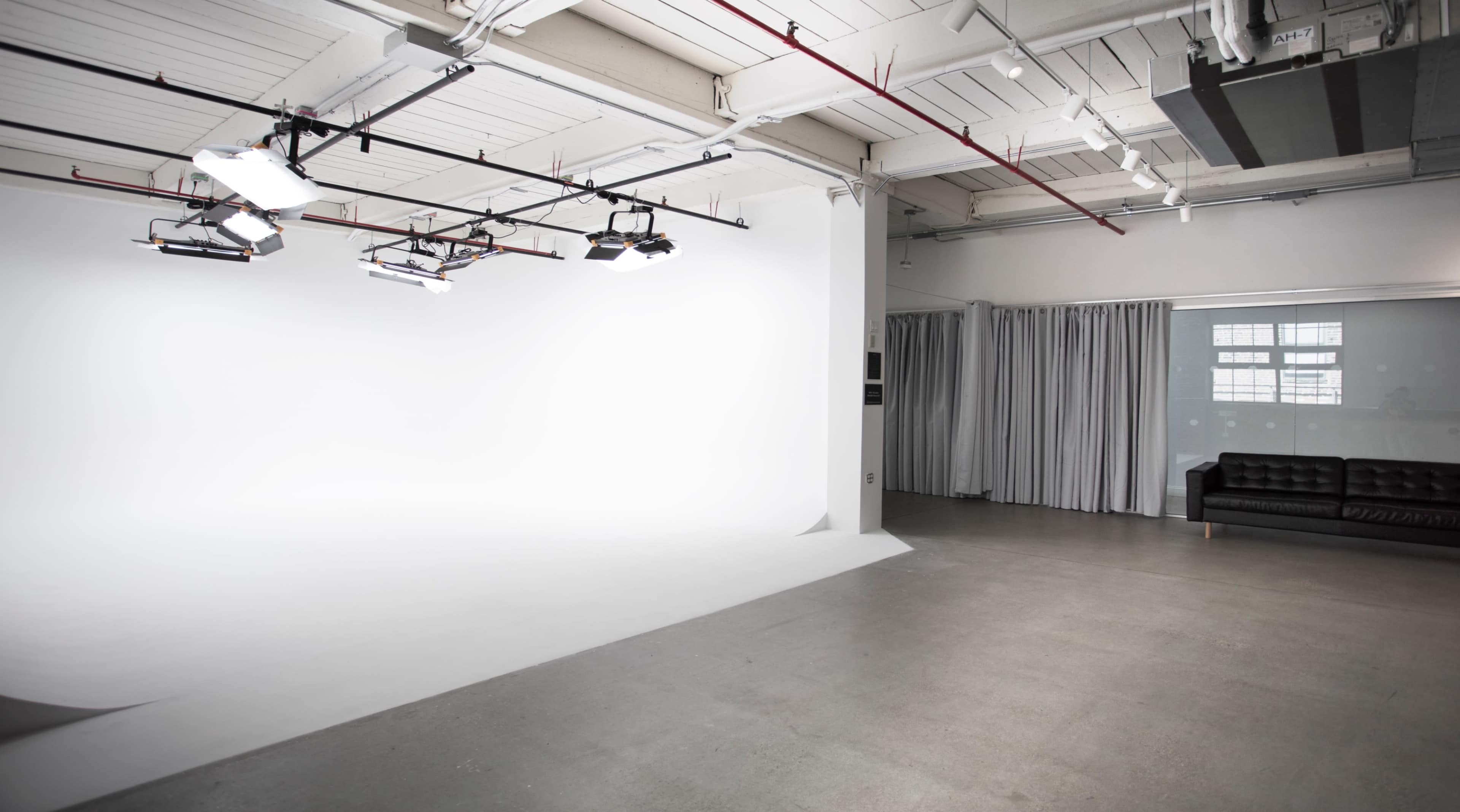 A spacious studio with a white backdrop, overhead lights, and a black sofa near a curtain.