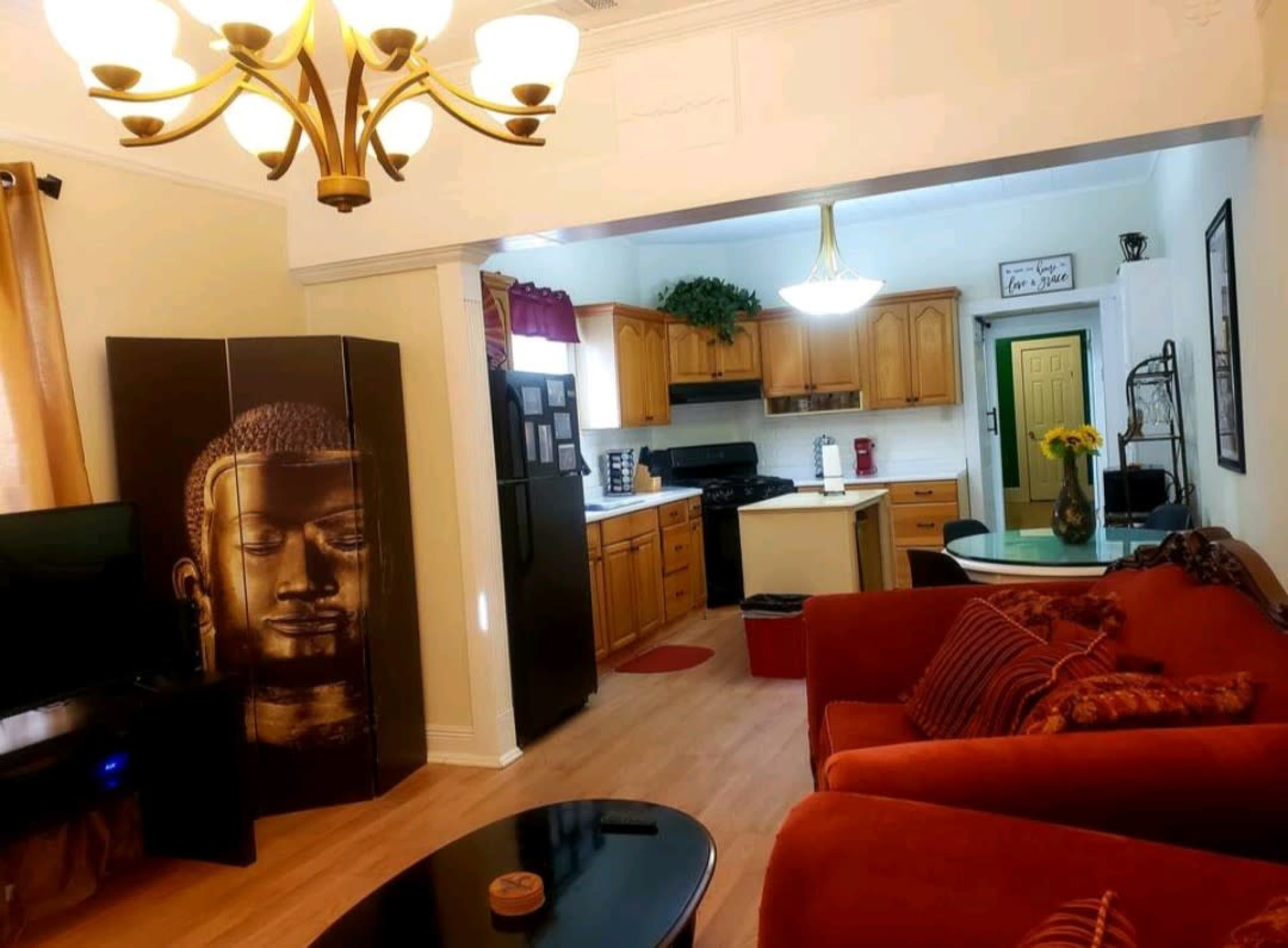 The image shows a living room area with a red couch, a black coffee table, a decorative screen featuring a Buddha face, and a kitchen visible in the background.