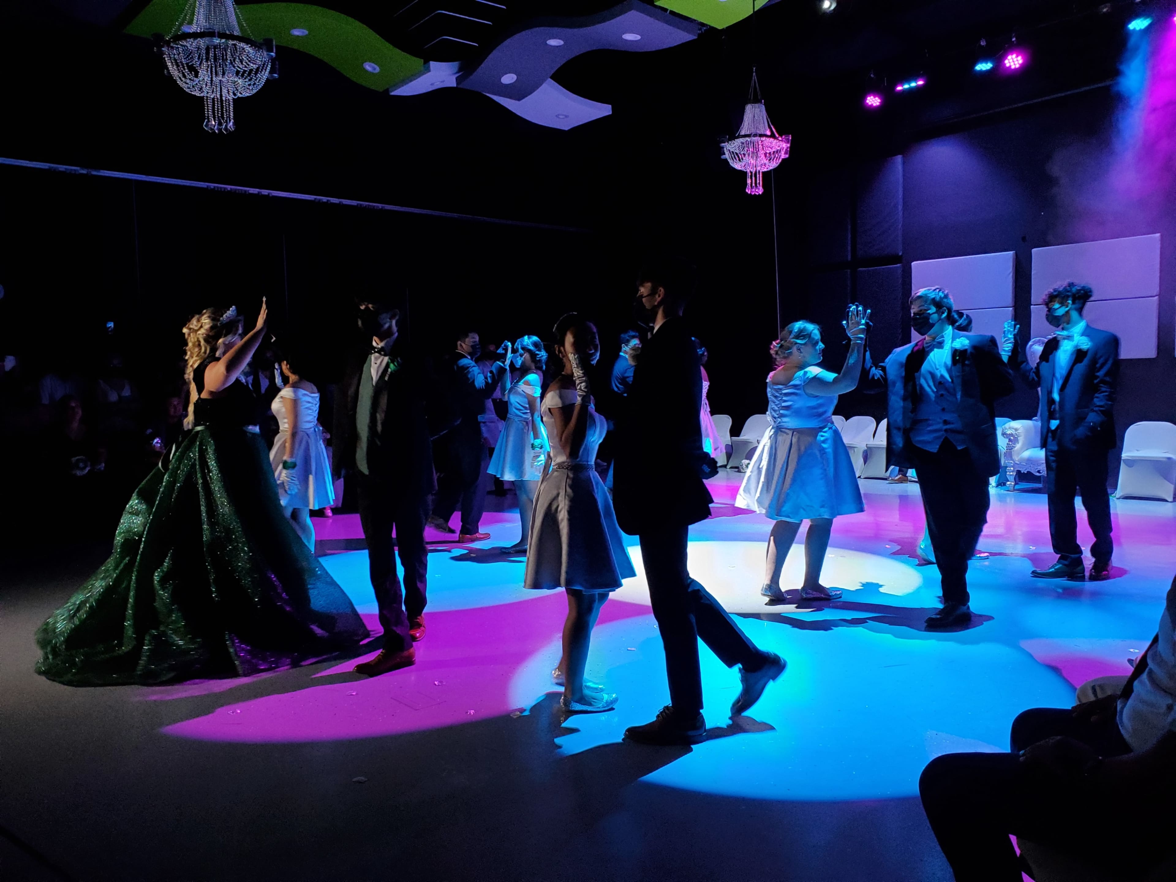 A group of people in formal attire dances on a stage illuminated by colorful lights, with chandeliers hanging above.