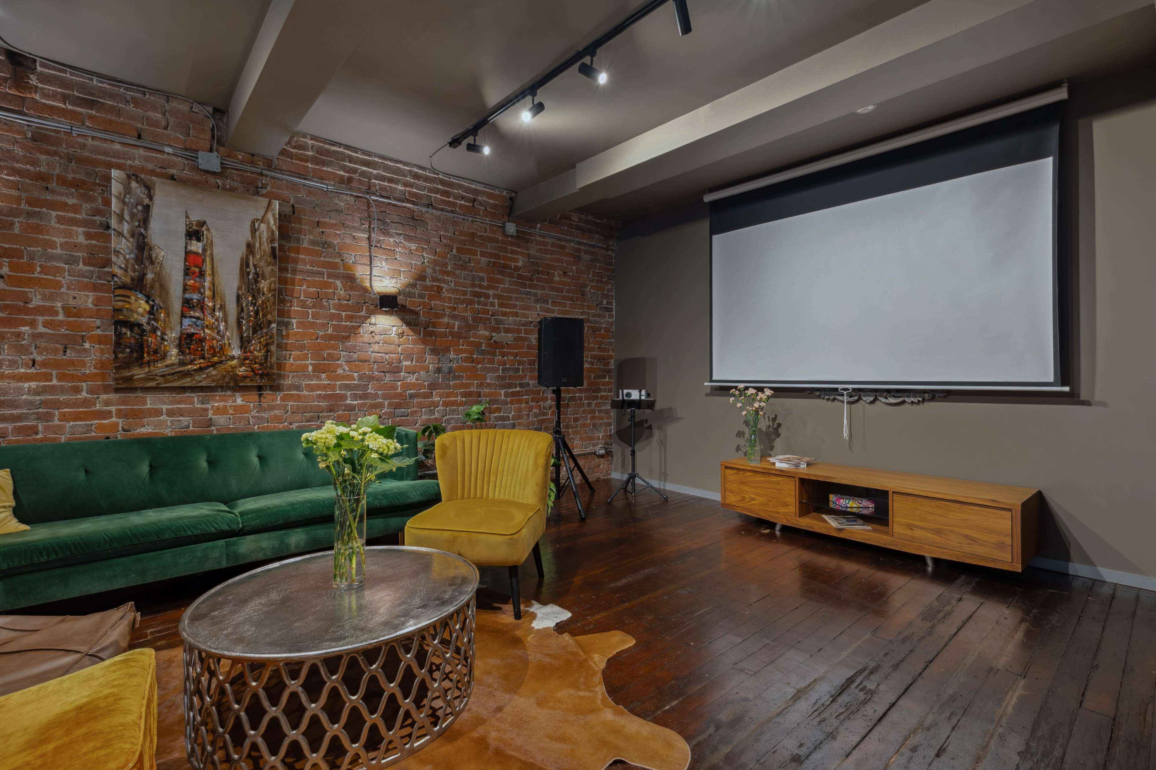 The image shows a modern living space featuring a green sofa, a yellow chair, a round coffee table, and a large wall projector screen, with exposed brick walls and hardwood flooring.