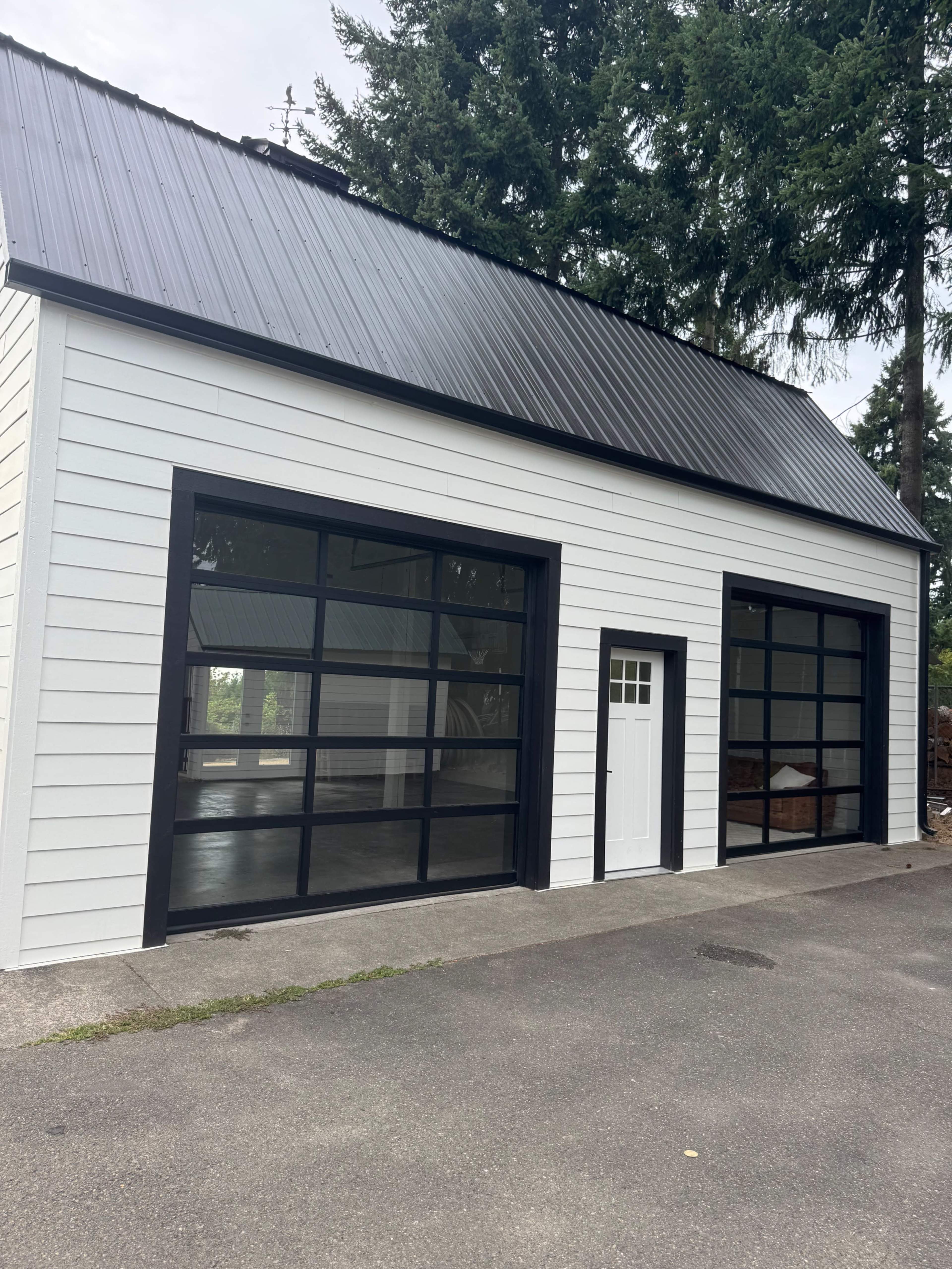 The image shows a modern white building with a black metal roof and large glass-paneled garage doors, situated on a paved driveway surrounded by trees.