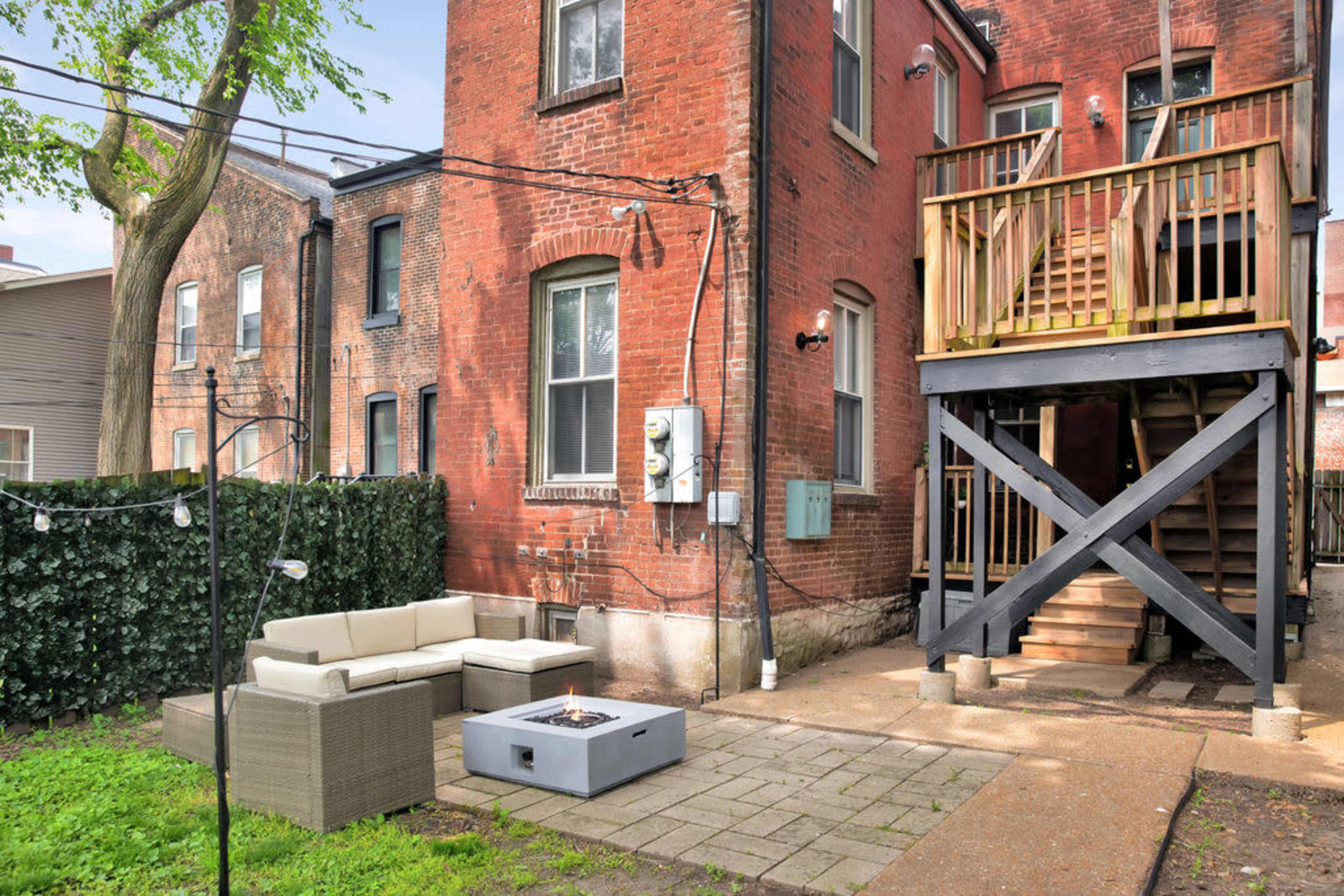 A backyard area with a seating arrangement and a fire pit near a brick building with a wooden staircase leading to an upper level.