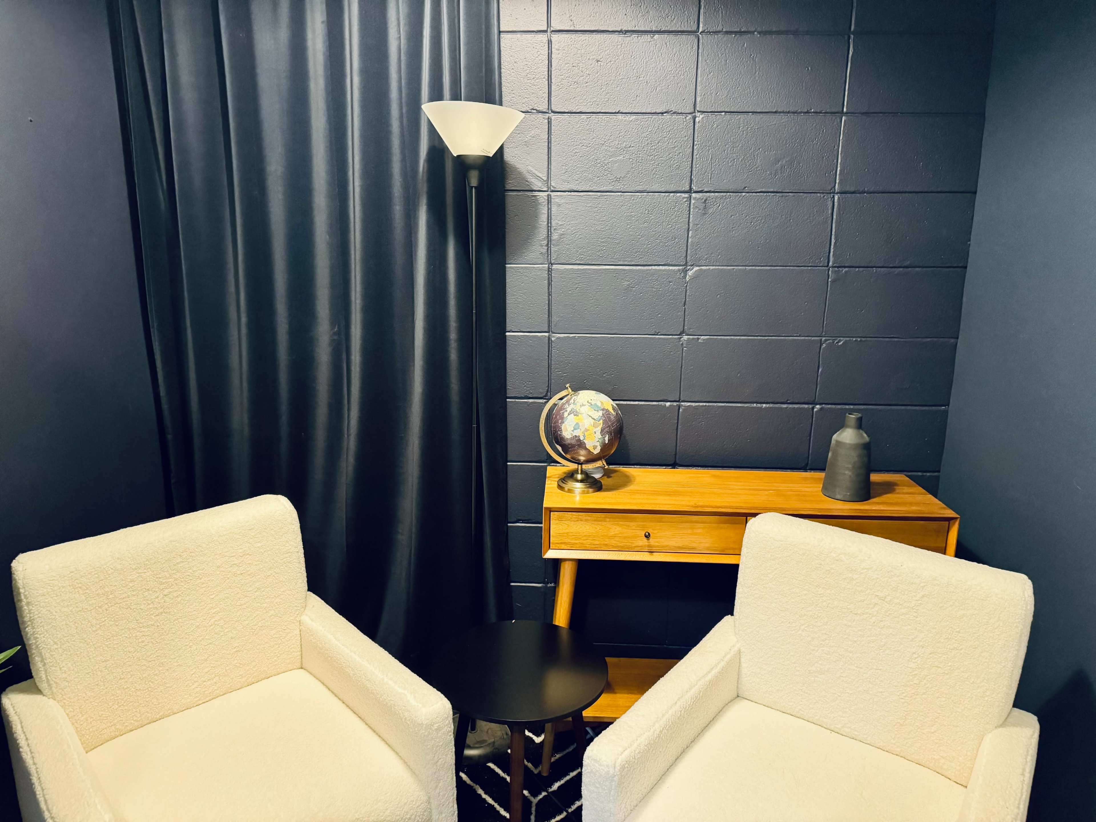 The image shows a cozy seating area with two white armchairs, a small black side table, a wooden console table, and a globe, all set against a dark blue wall and a curtain.