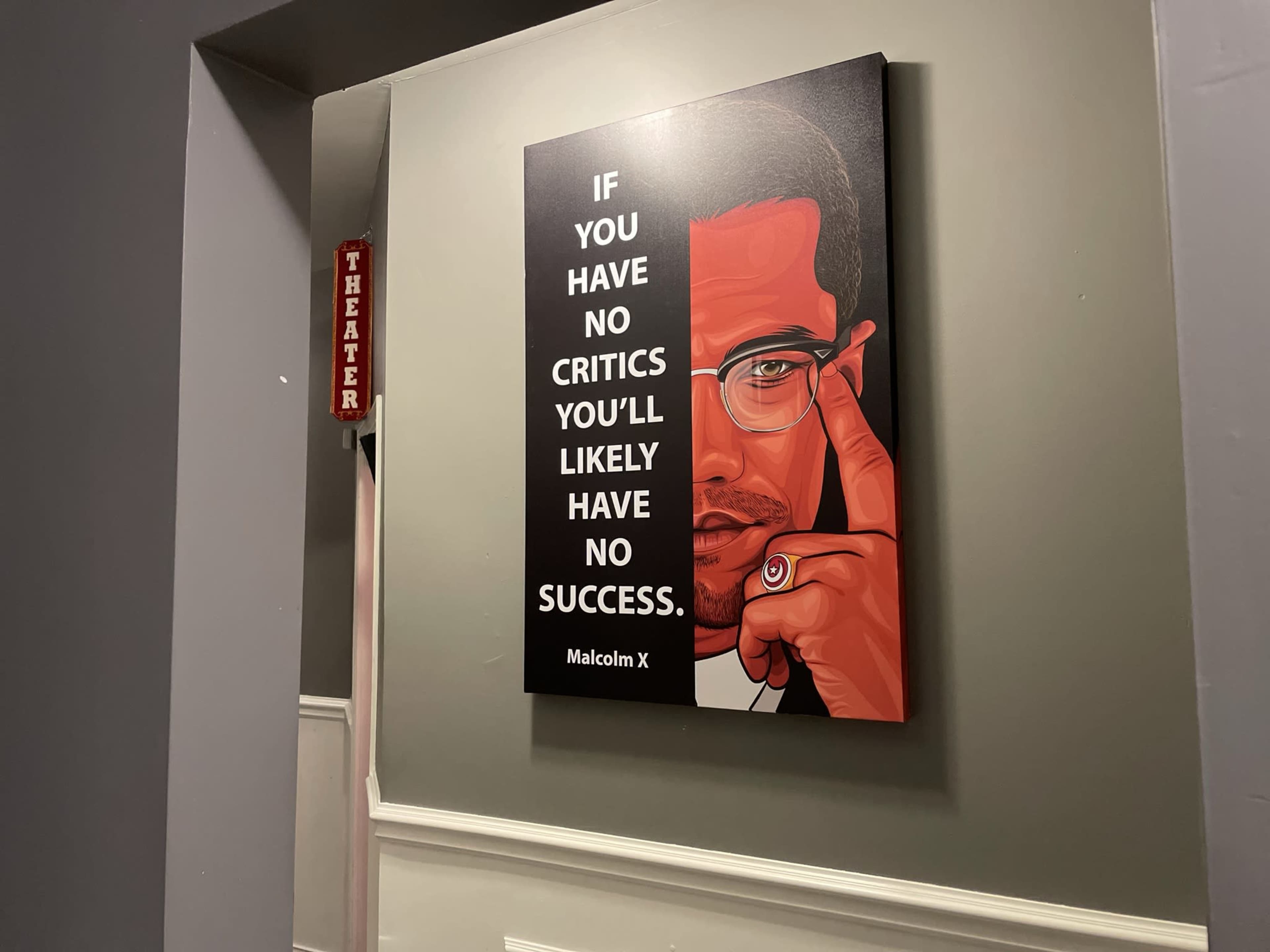 A poster of Malcolm X with the quote, "IF YOU HAVE NO CRITICS YOU'LL LIKELY HAVE NO SUCCESS," hangs on a wall next to a sign labeled "THEATER."