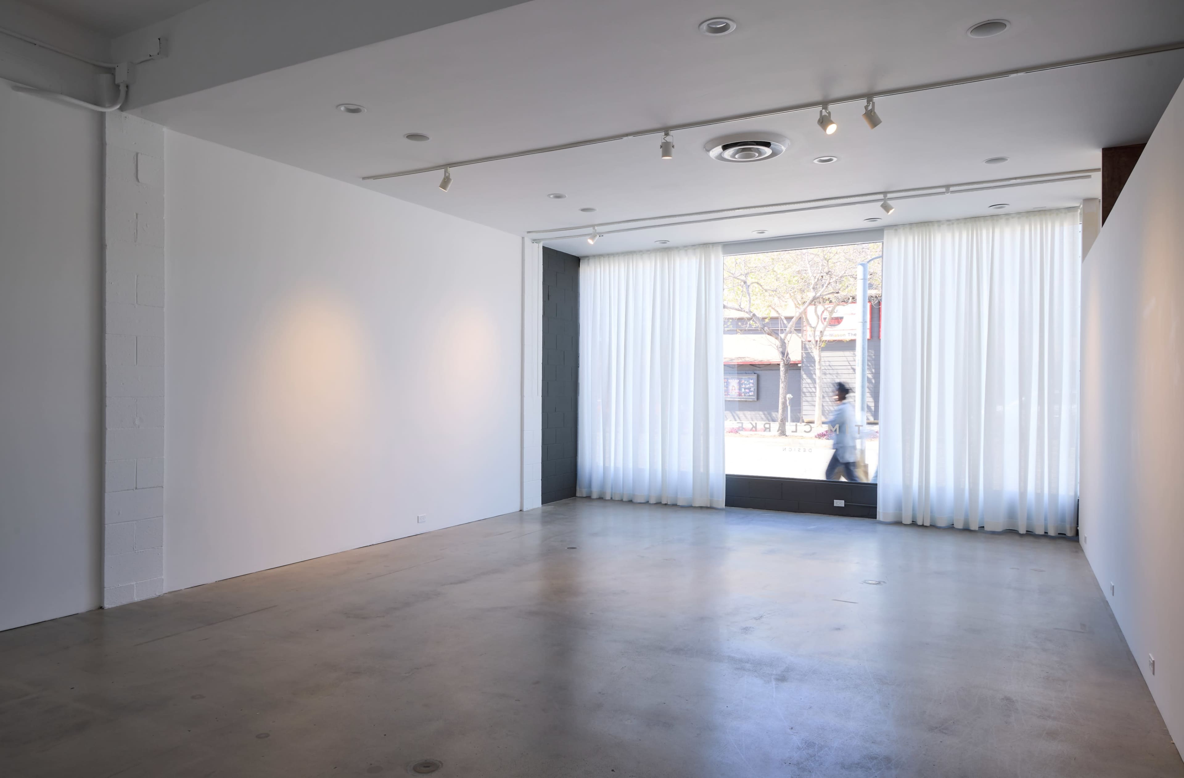 The image shows a spacious, empty gallery with white walls and large windows covered by sheer curtains.