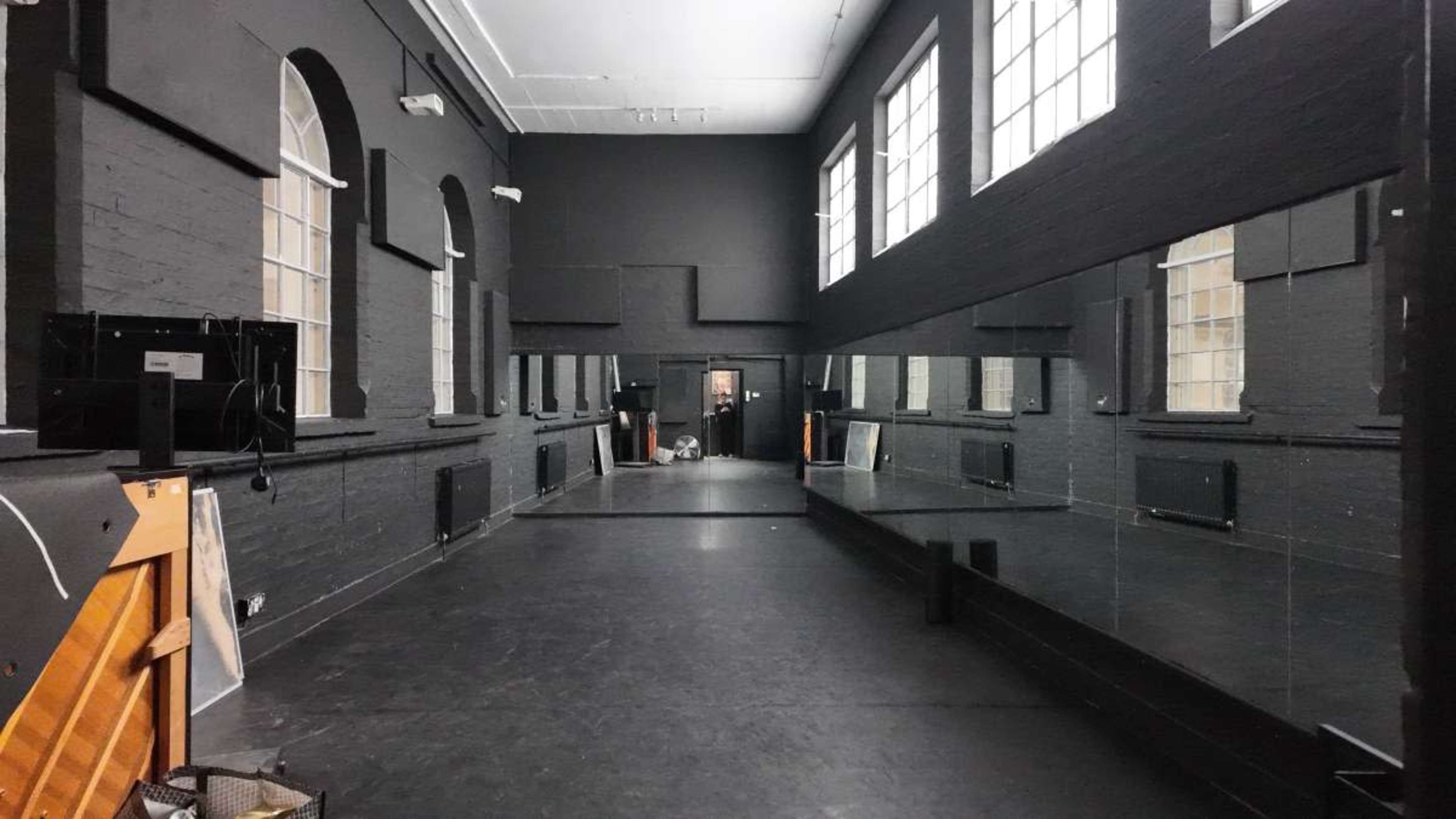 The image shows a long, narrow room with black walls, large windows, and mirrors lining one side, along with a piano in the foreground.