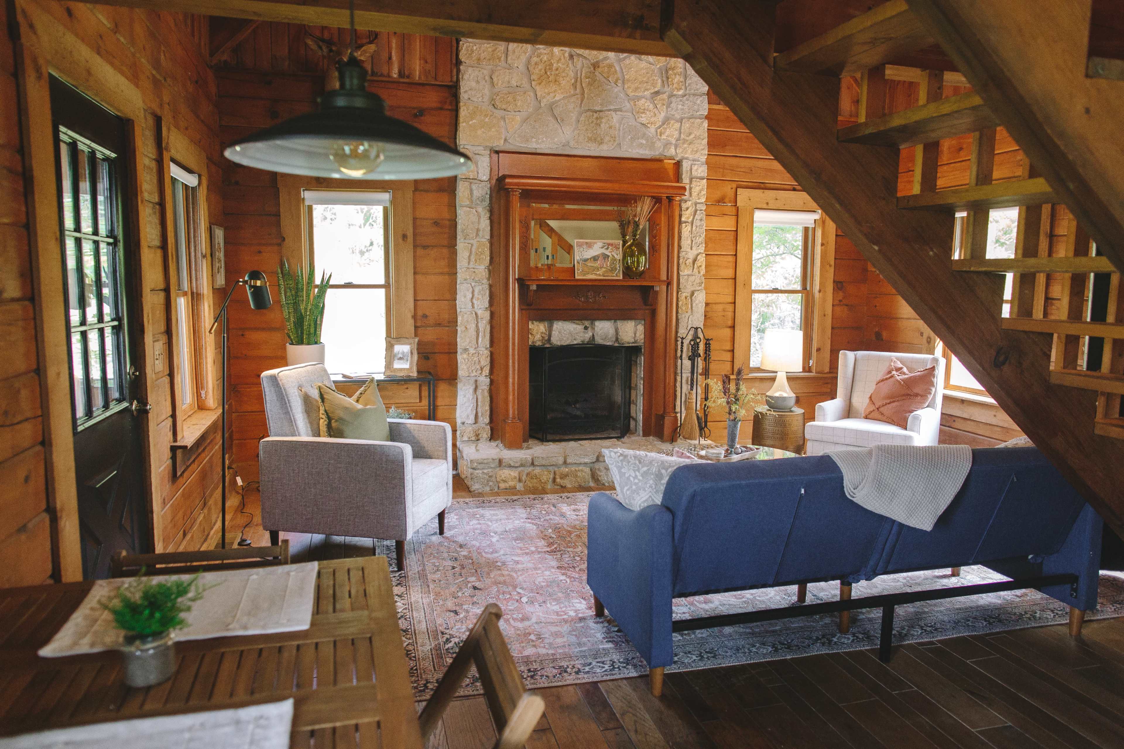 A cozy, wooden cabin interior featuring a living area with two chairs and a blue sofa arranged around a stone fireplace.