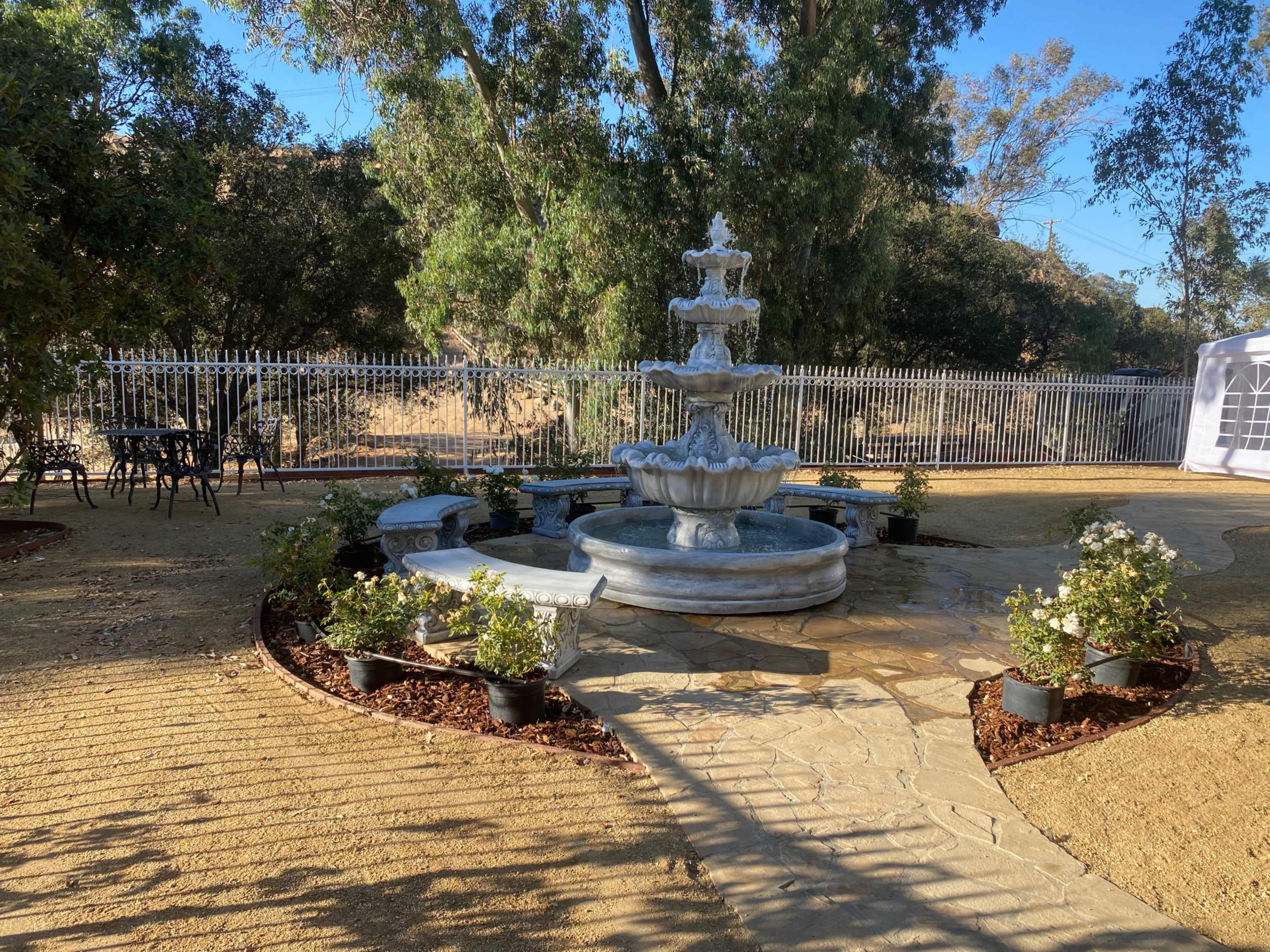 A three-tiered stone fountain is surrounded by potted plants and a stone pathway in a landscaped outdoor area.
