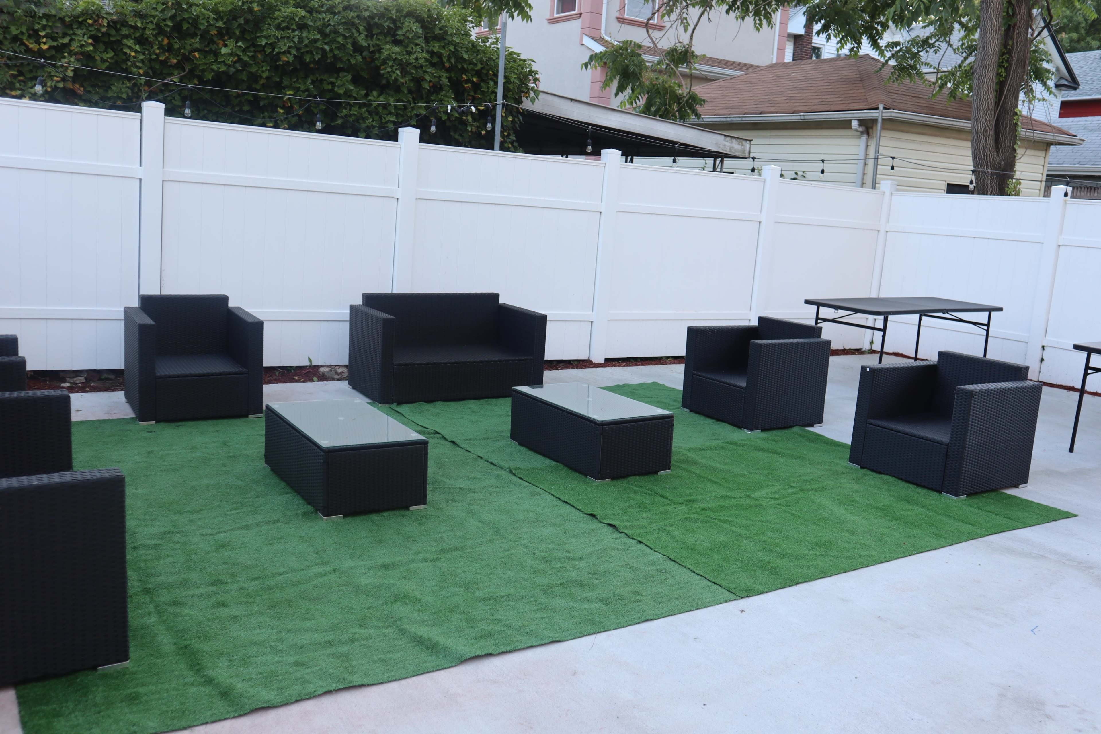 A patio area features a set of black wicker furniture, including a sofa, chairs, and a glass-topped coffee table, arranged on green artificial grass.