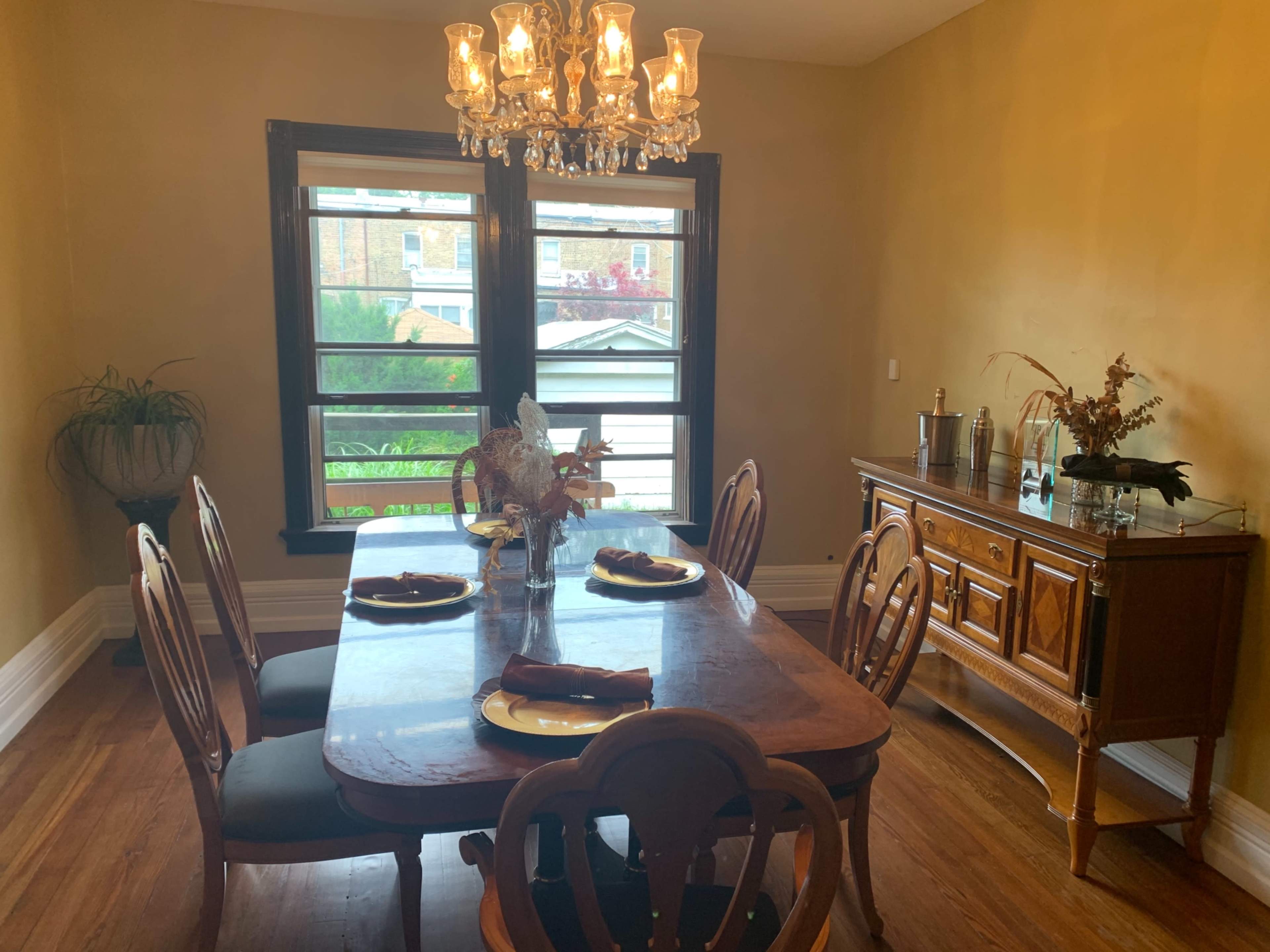 A dining room features a wooden table set for four, with a chandelier above and a sideboard against the wall.