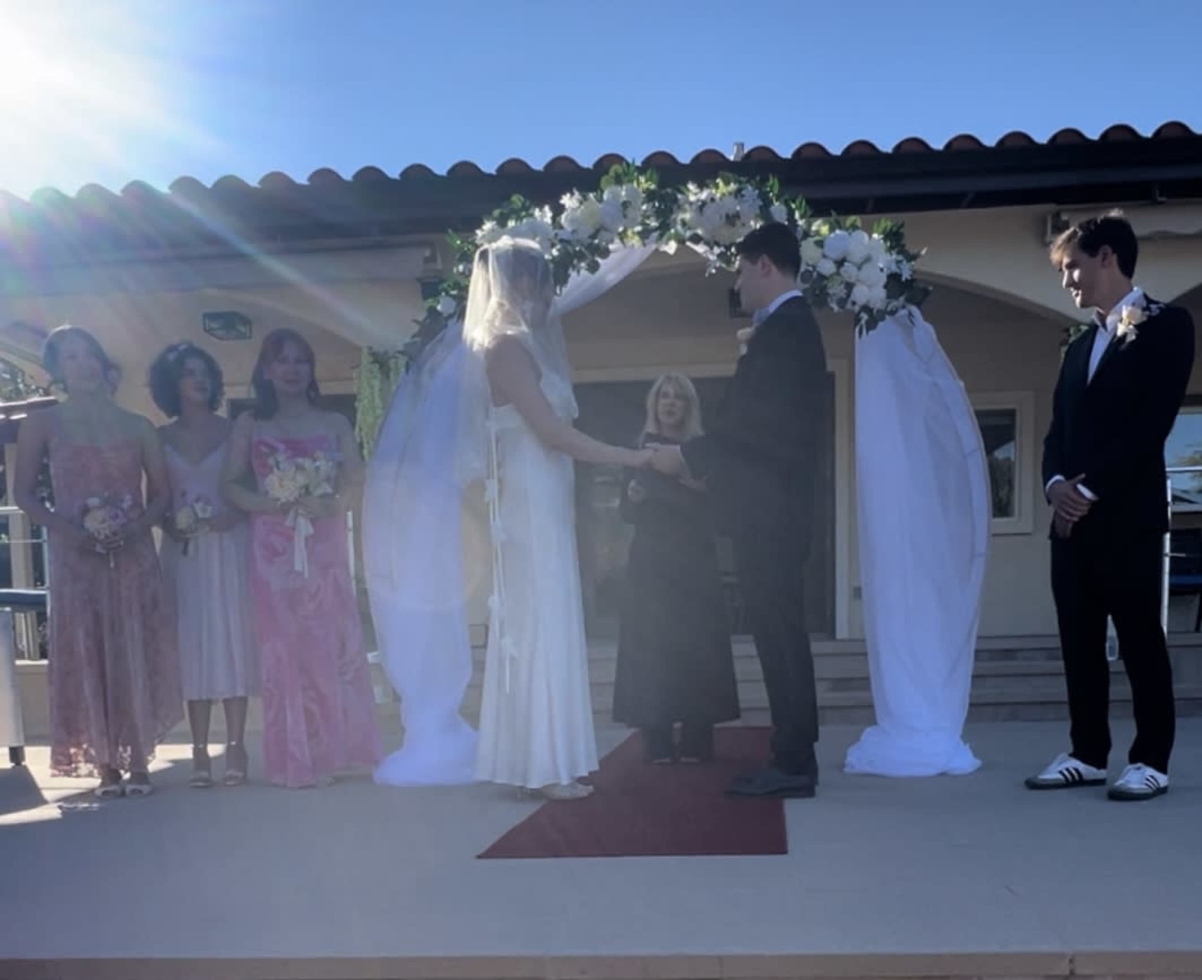 A couple stands under a floral arch, exchanging vows while surrounded by their wedding party on an outdoor stage.