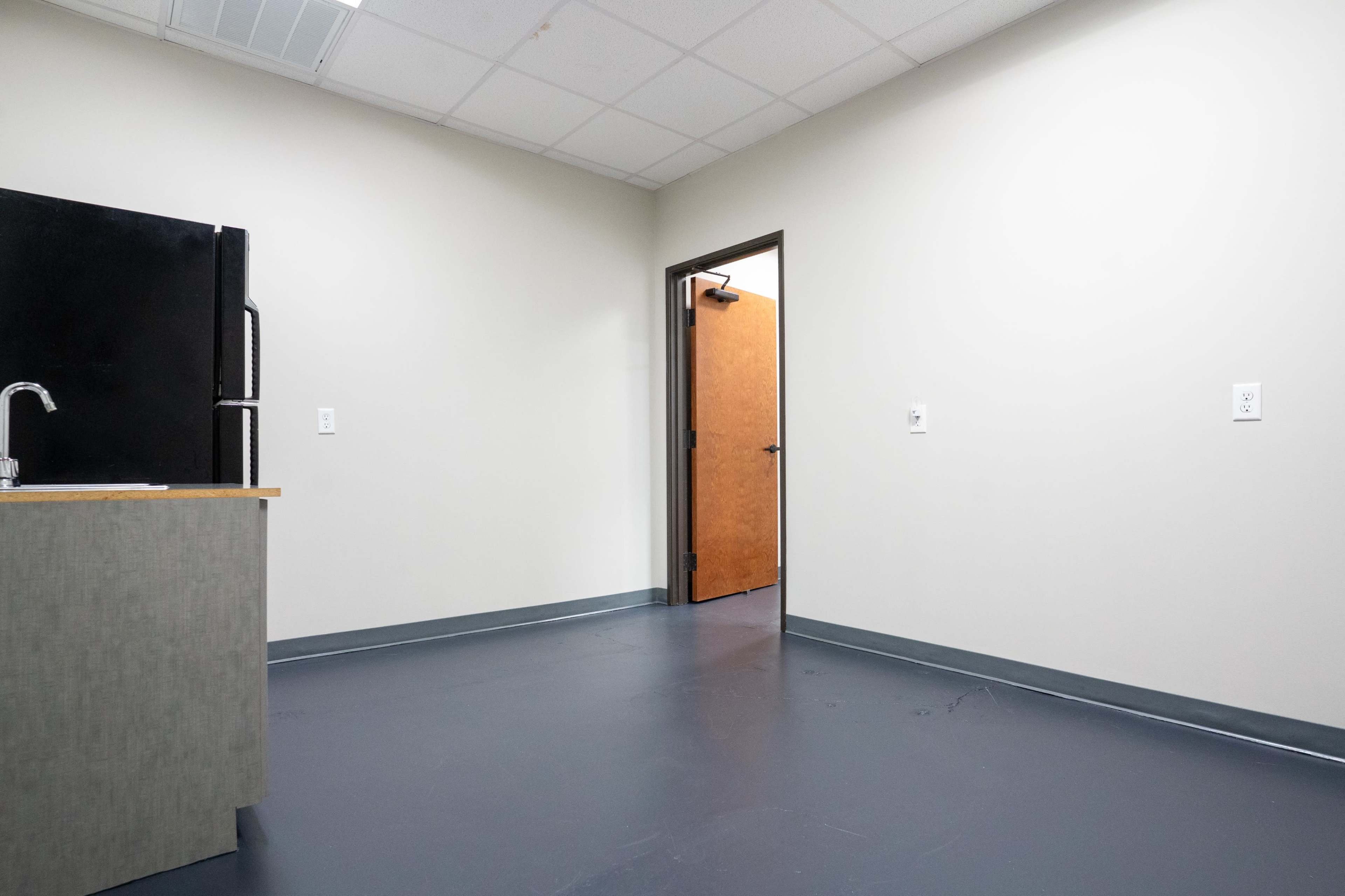 The image shows an empty room with a kitchen area on the left and a wooden door on the right leading to another space.