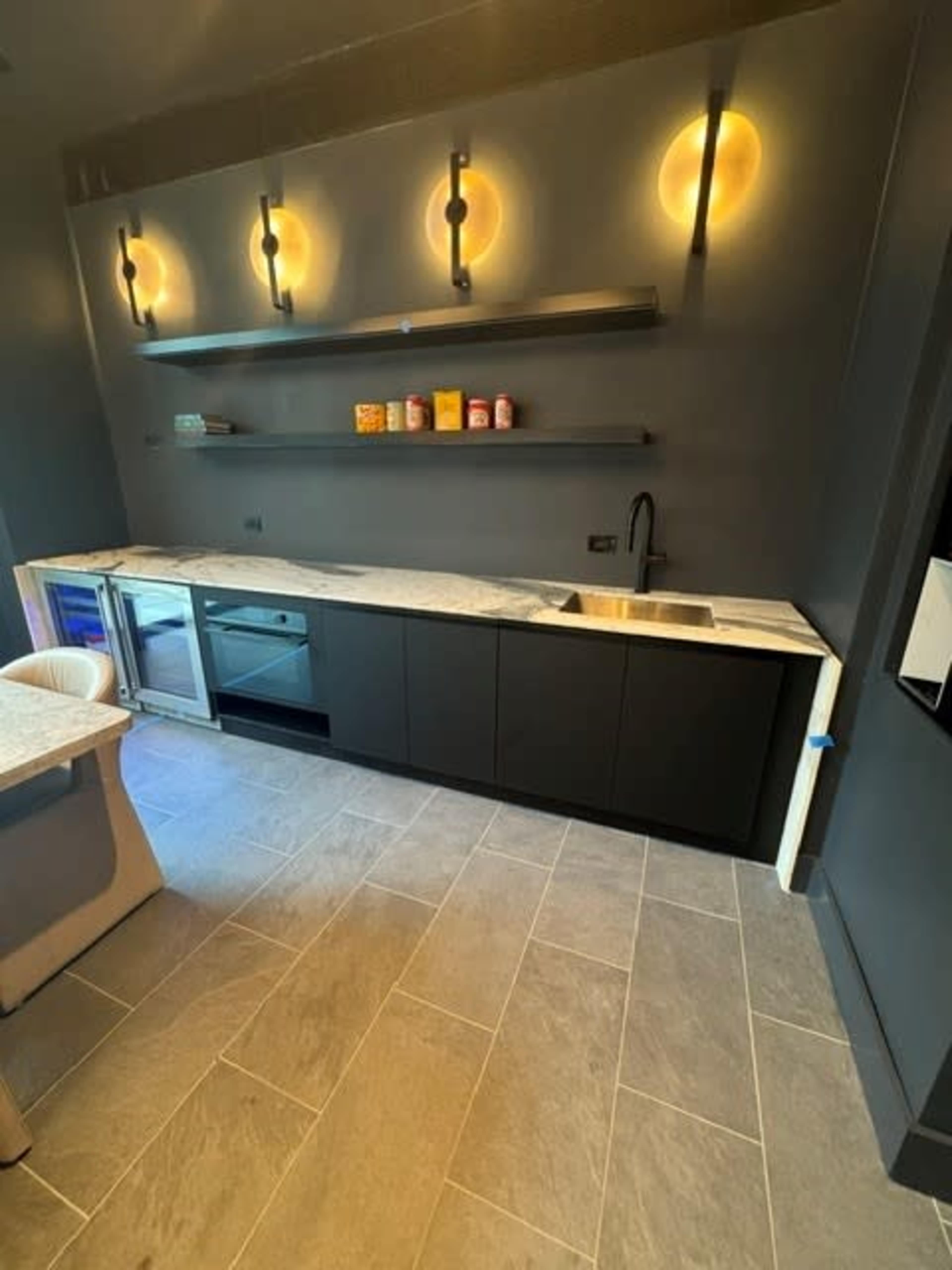 The image shows a modern kitchen with dark cabinetry, a light-colored countertop, and minimalistic shelves illuminated by circular lights.