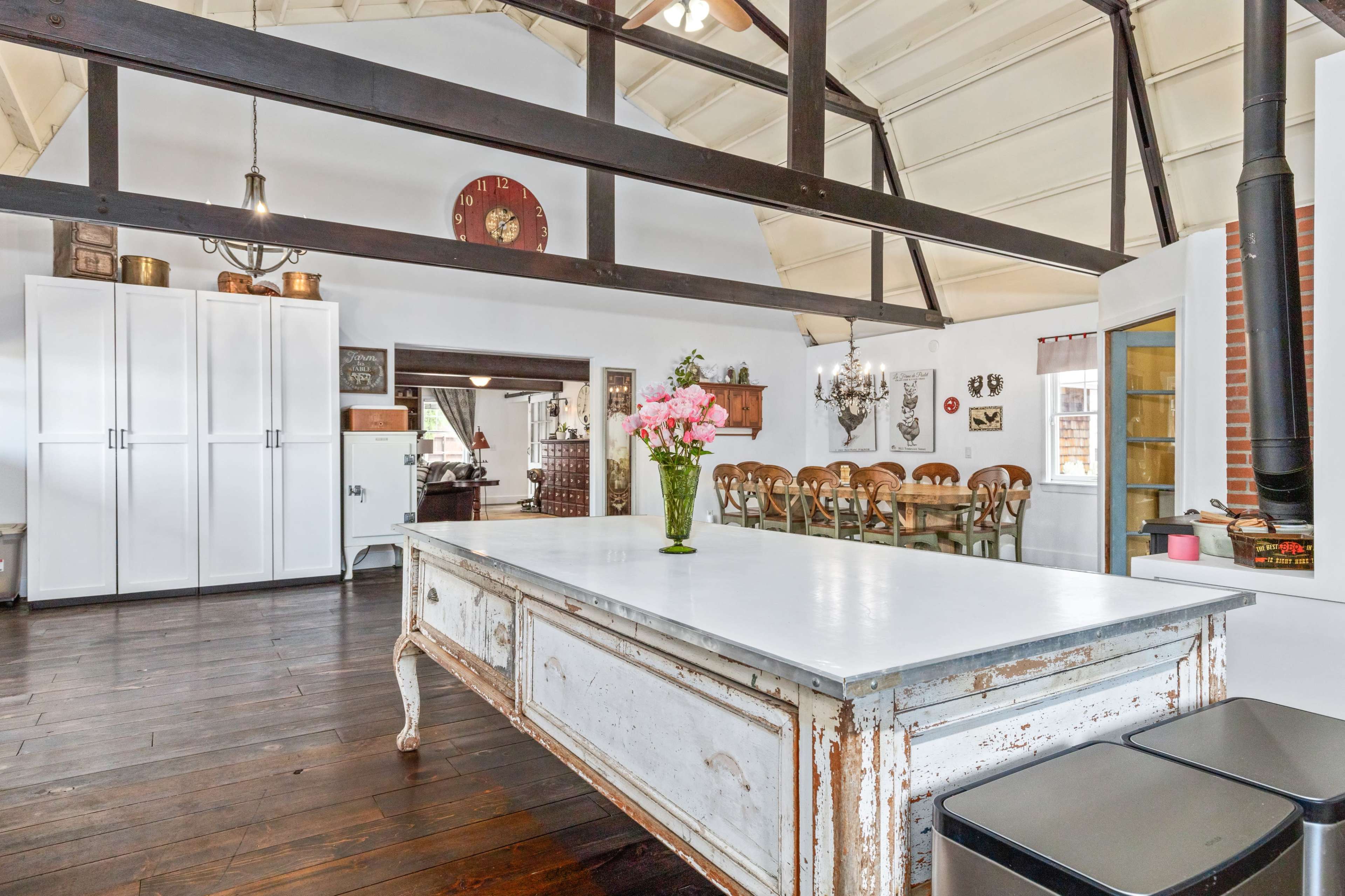 A spacious kitchen features a large white table in the center, surrounded by dark wooden floors and exposed beams overhead.