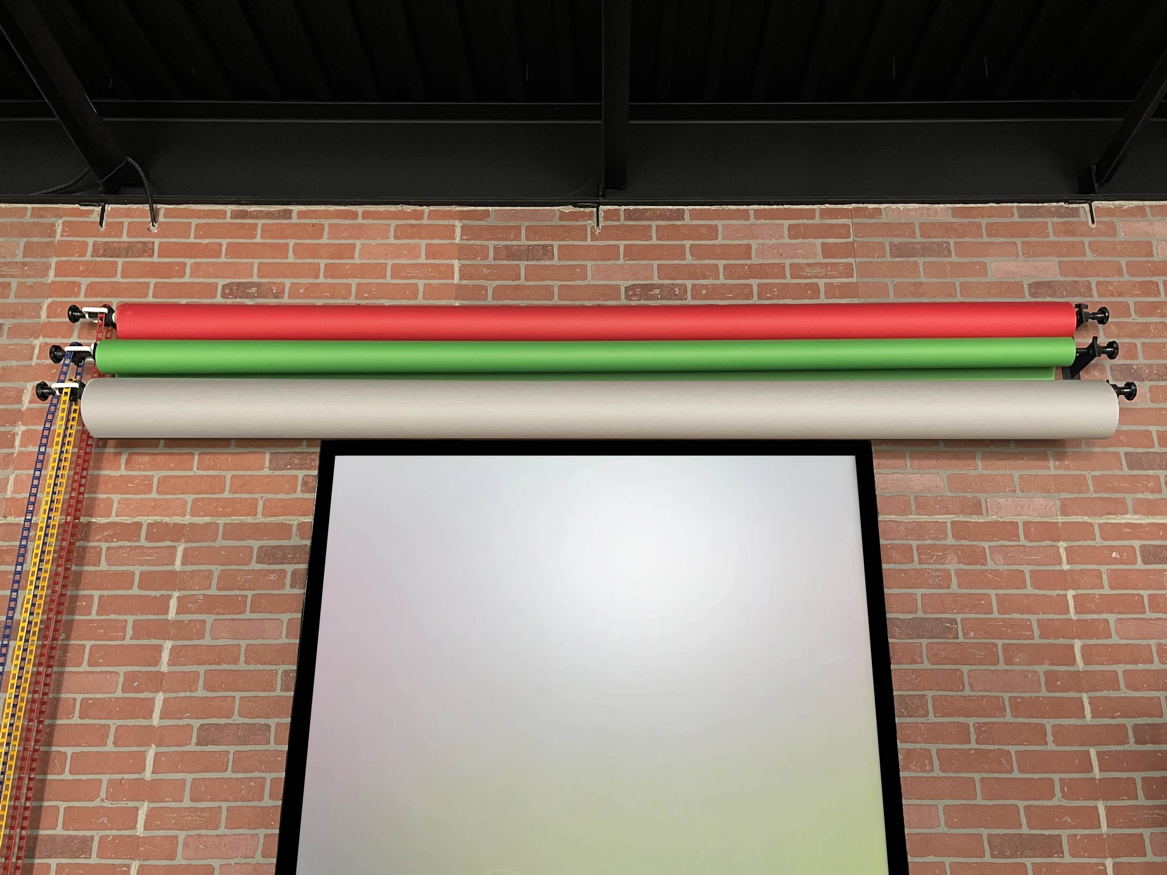 The image shows a brick wall with three rolls of colored paper—red, green, and a beige—mounted above a large projection screen.
