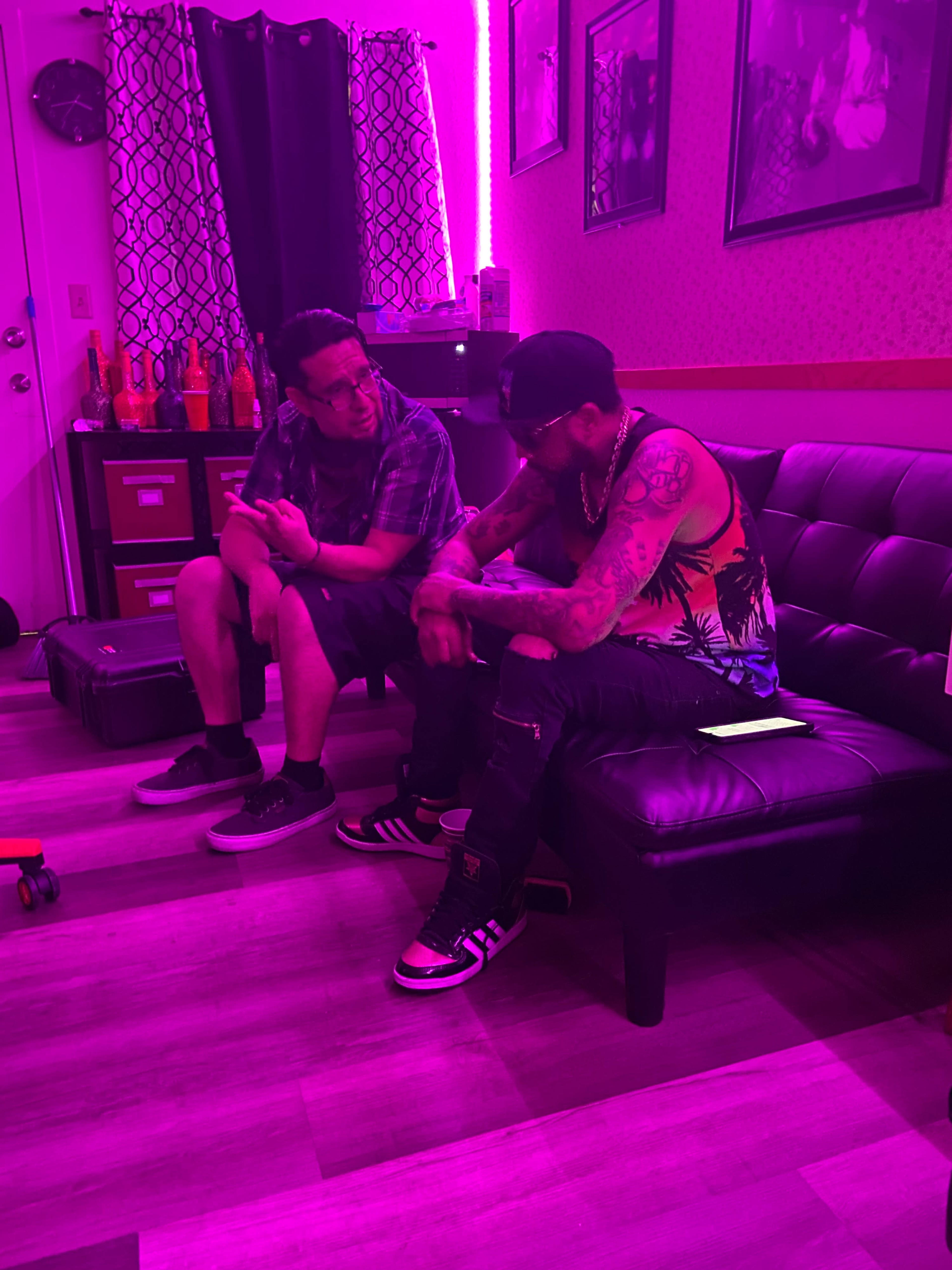 Two people are seated on a black couch in a room illuminated by pink lights, engaged in conversation.
