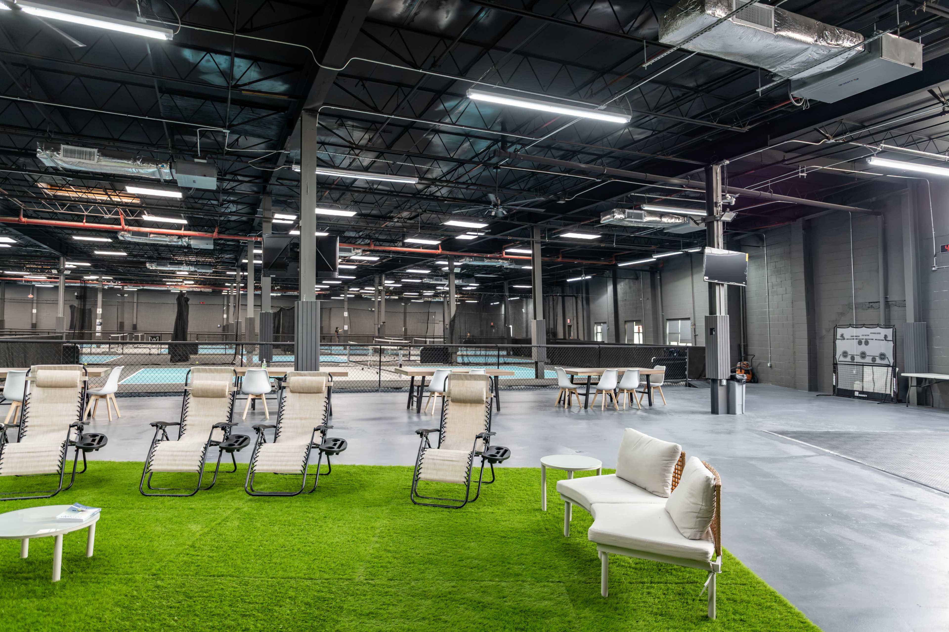 The image shows an expansive indoor recreational space featuring a seating area on artificial grass with lounge chairs and tables, alongside a court area visible in the background.