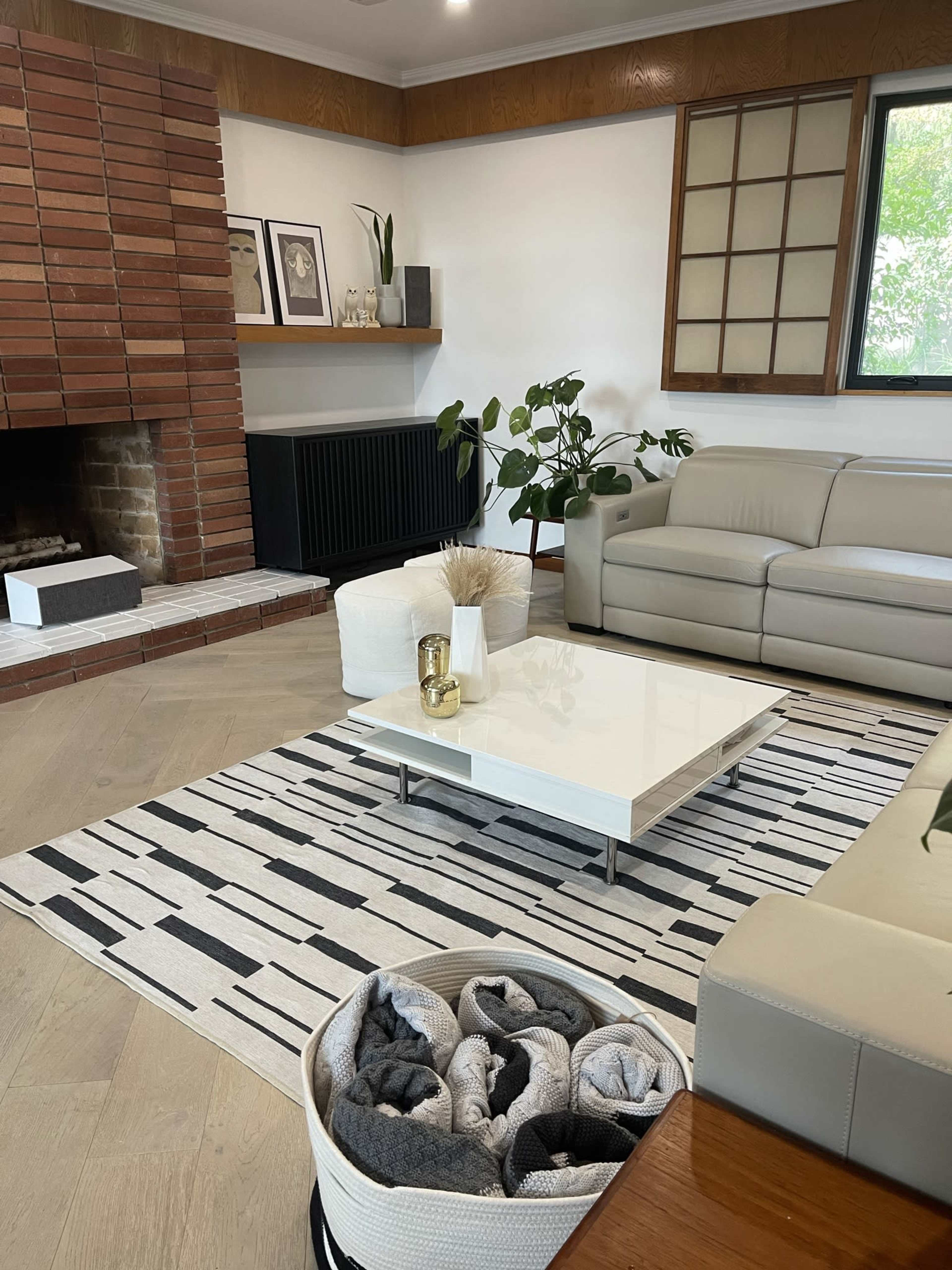 A modern living room with a white coffee table on a striped rug, a light-colored sofa, a brick fireplace, and plants arranged around the space.