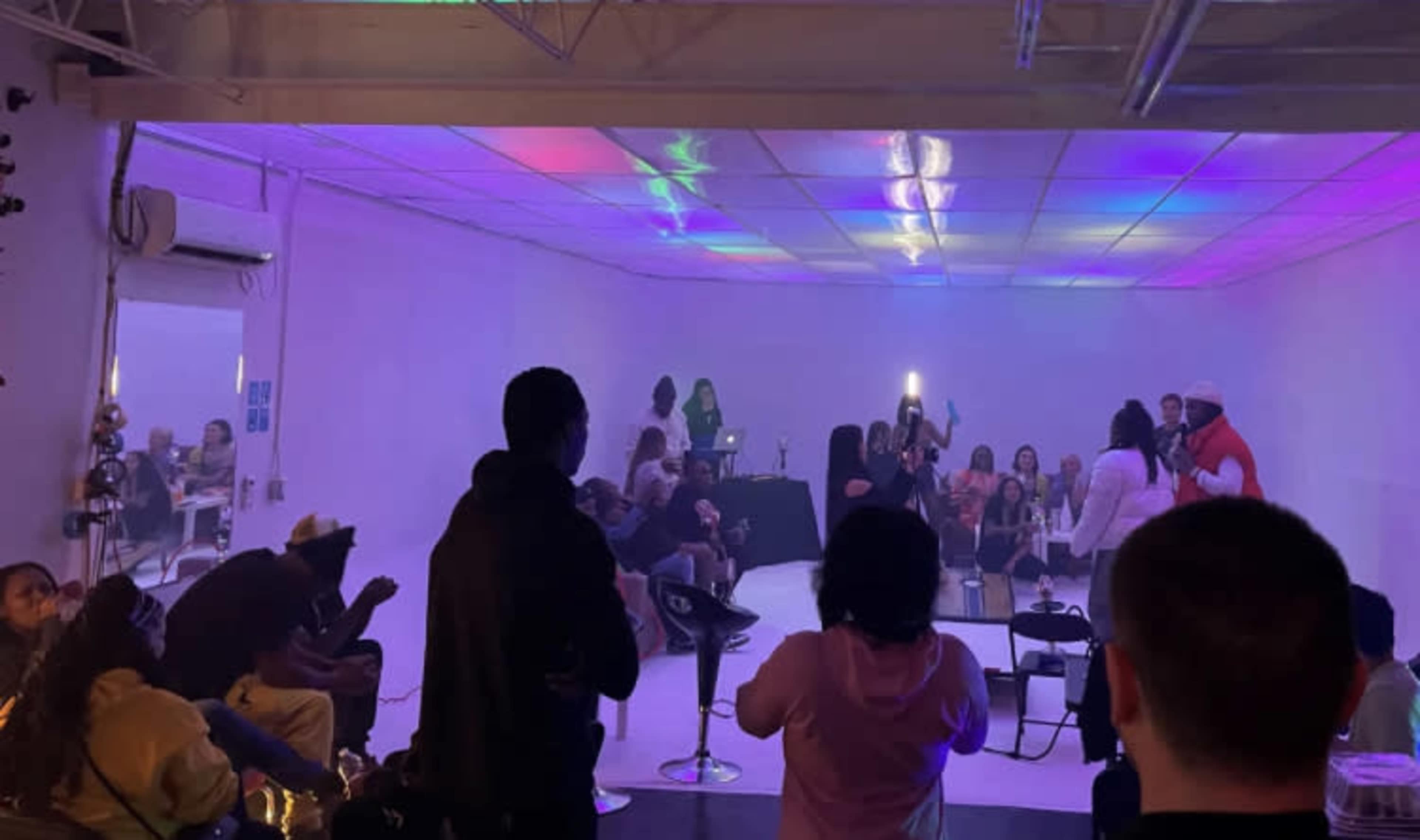 A group of people is gathered in a brightly lit room with colorful lights and chairs, while some individuals appear to be performing or presenting on a small stage.