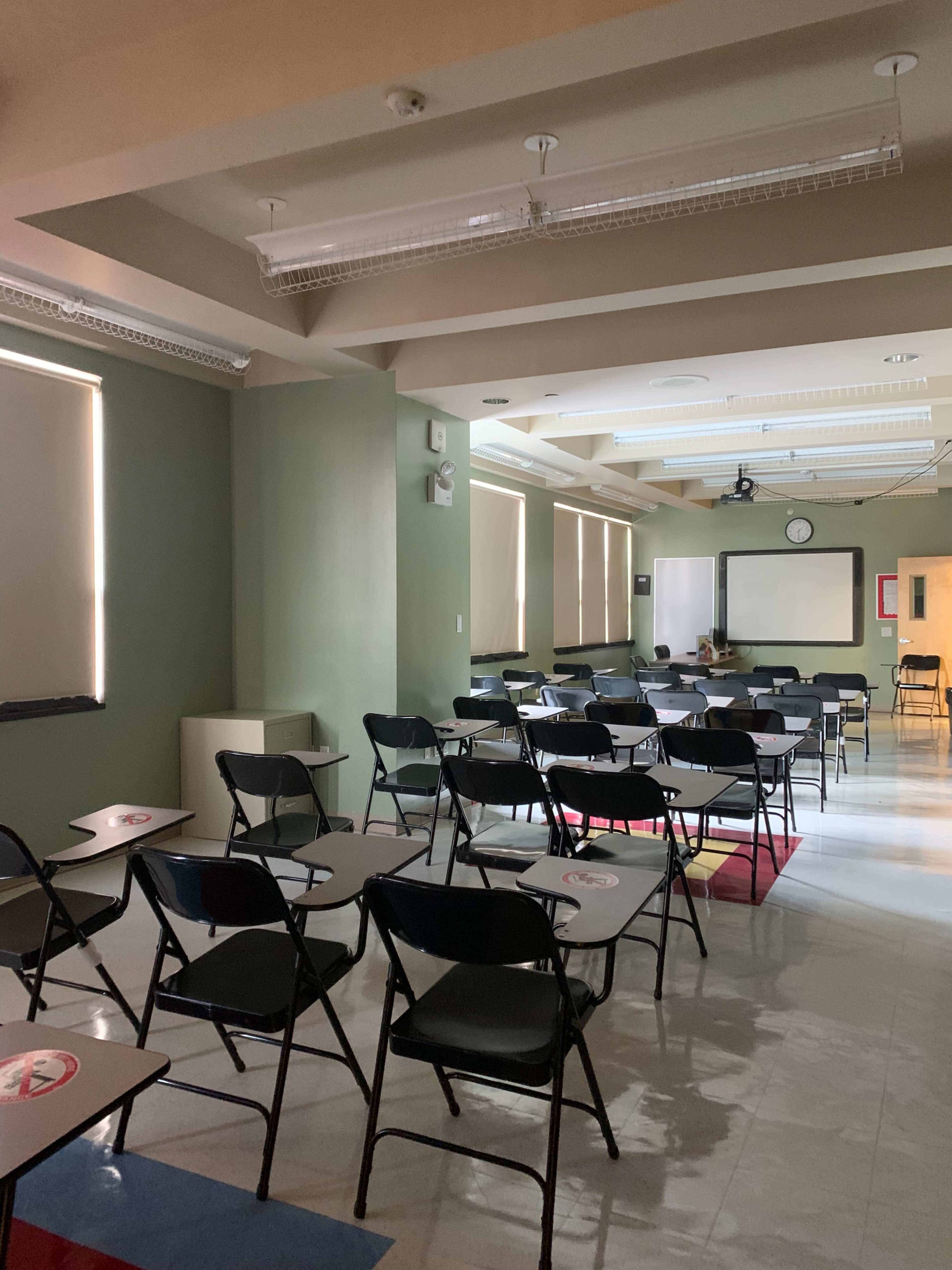 The image shows a classroom arranged with rows of black folding chairs and desks, illuminated by natural light from windows.