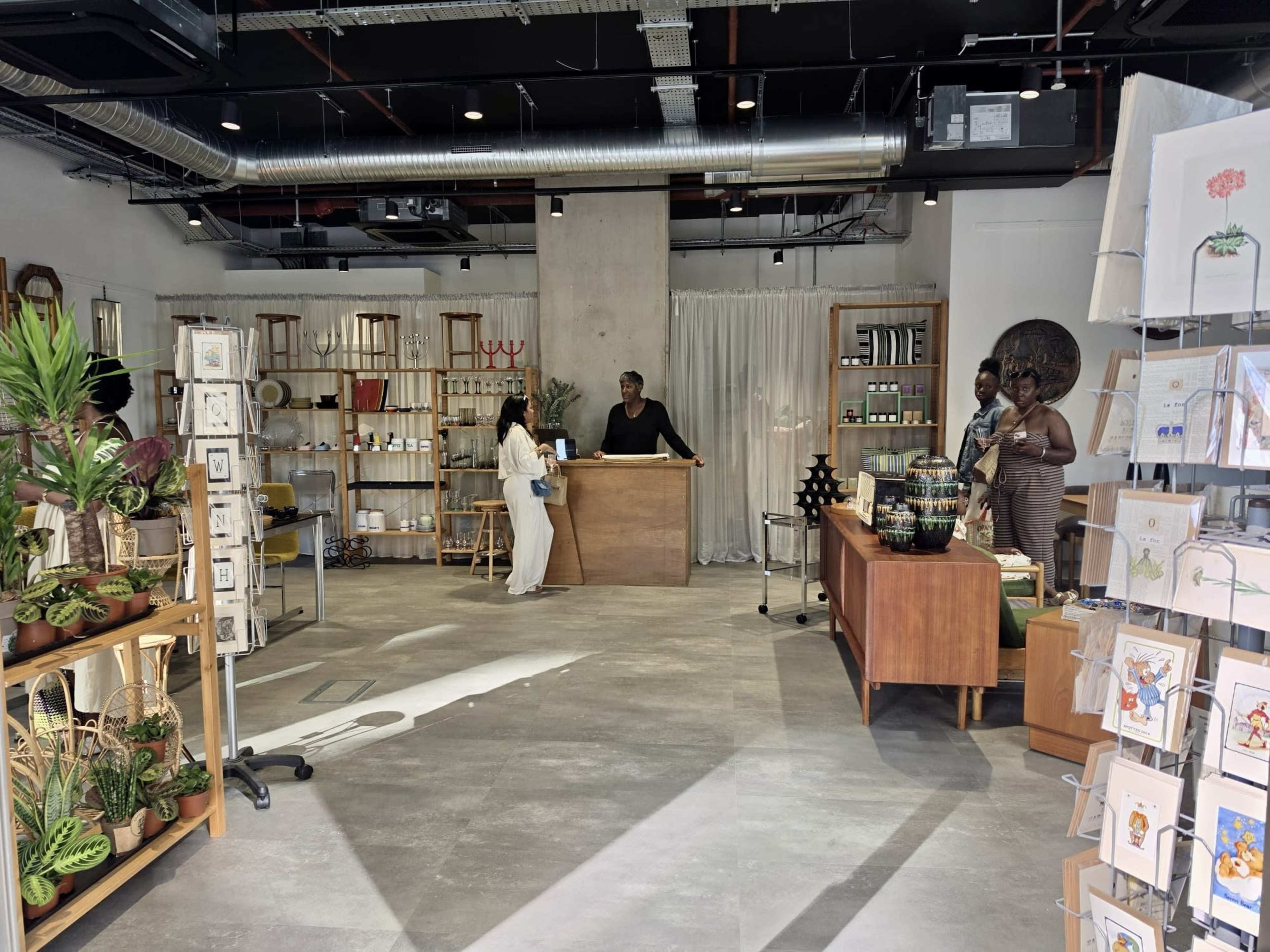 The image shows a well-organized retail space featuring a wooden checkout counter, shelves filled with products, and two customers interacting with a staff member.