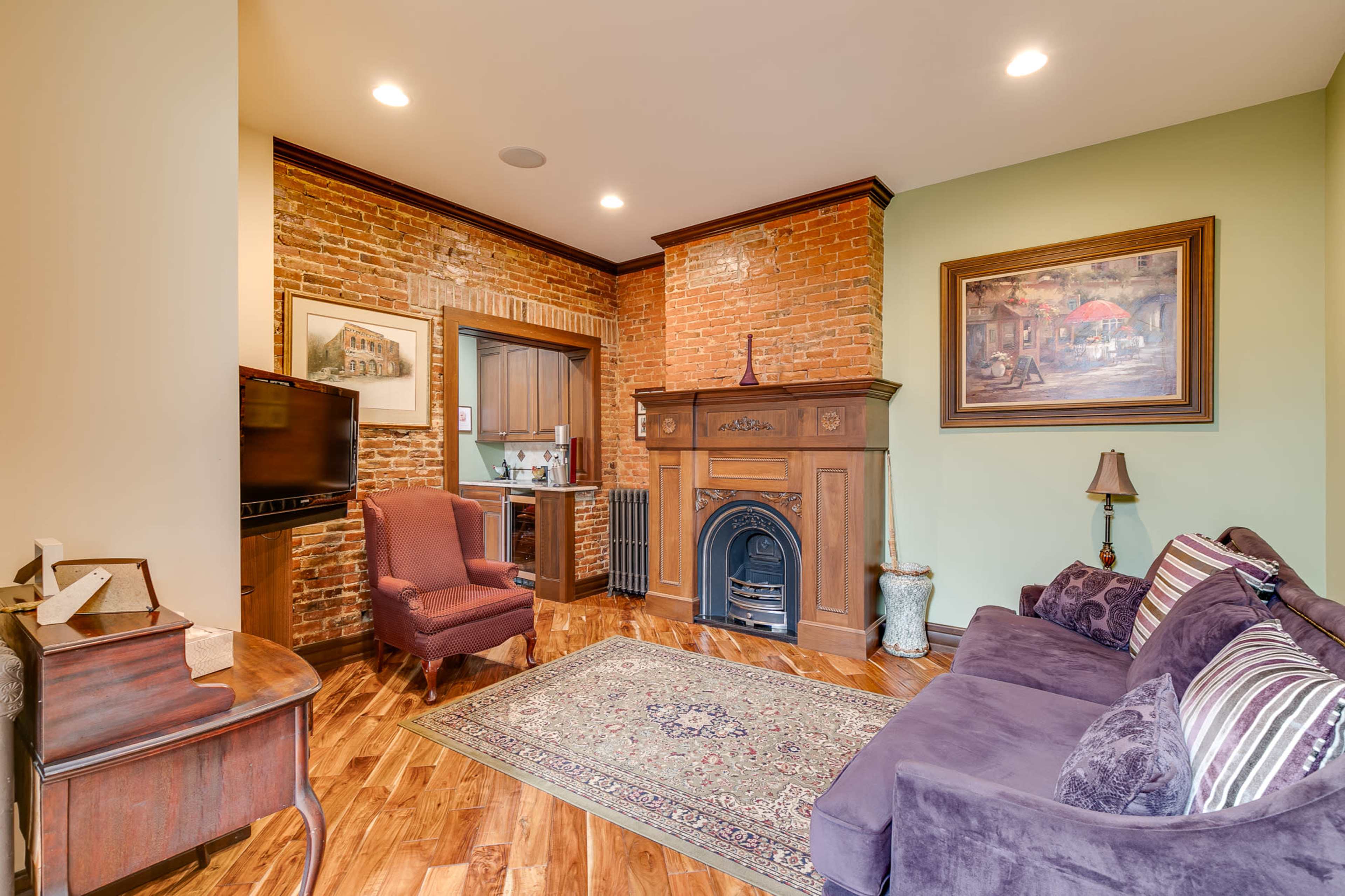 The image shows a cozy living room featuring a brick accent wall, a wooden fireplace, a purple sofa, and a wooden chair, with a decorative rug on the floor.