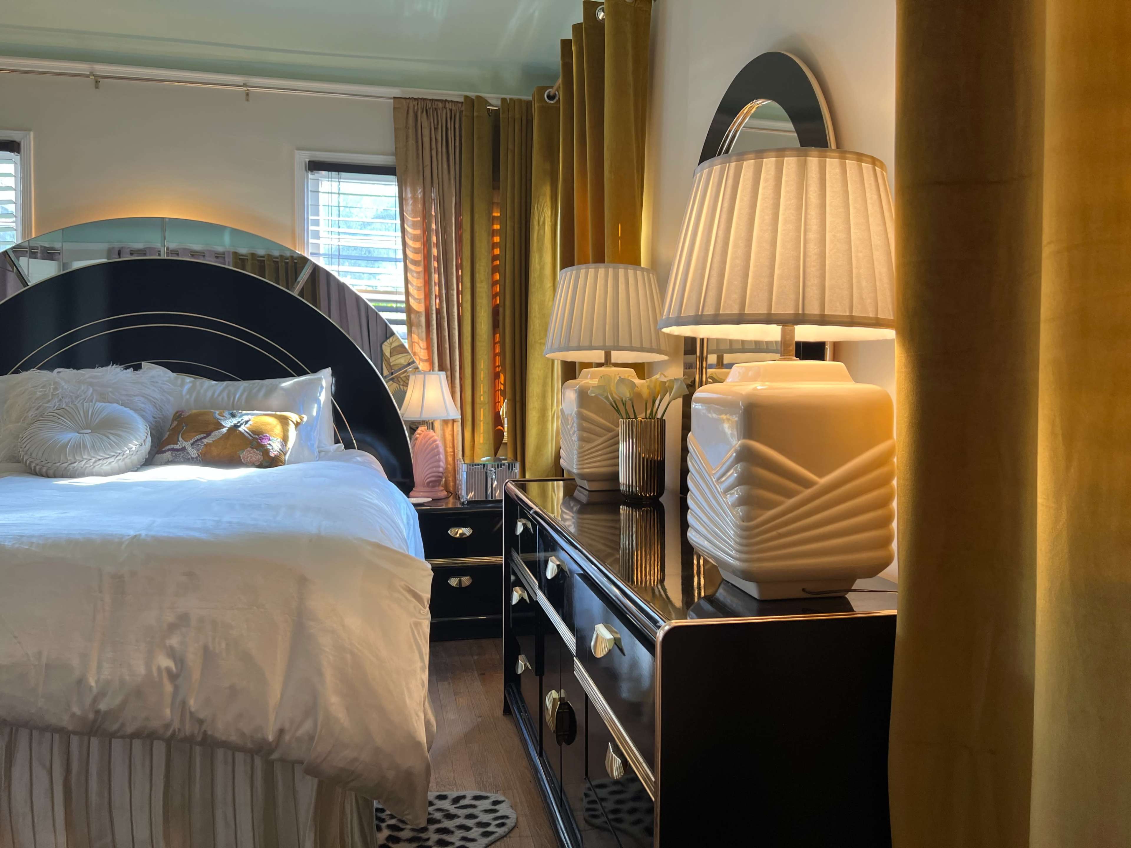 A well-decorated bedroom features a large bed with a curved headboard, two bedside lamps on a black dresser, and elegant curtains.