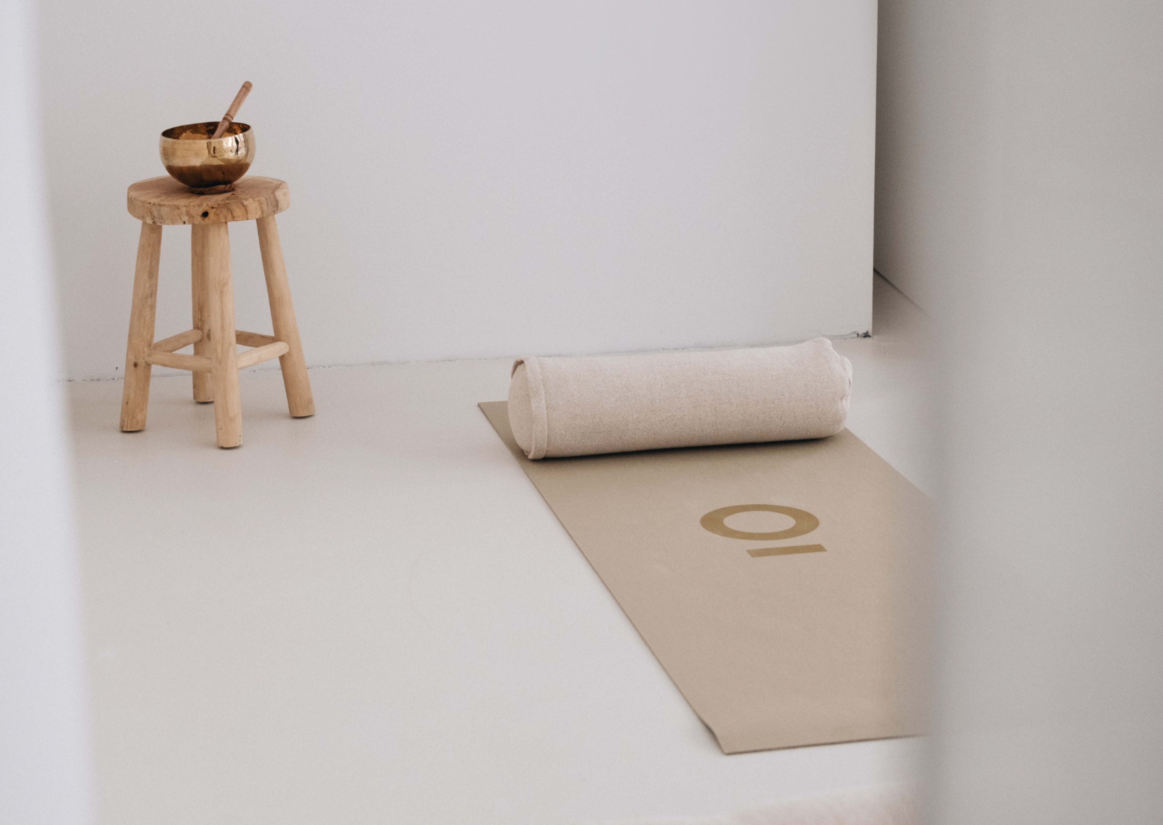 A rolled yoga mat with the number "10" is positioned next to a wooden stool and a metal bowl in a minimalistic room.
