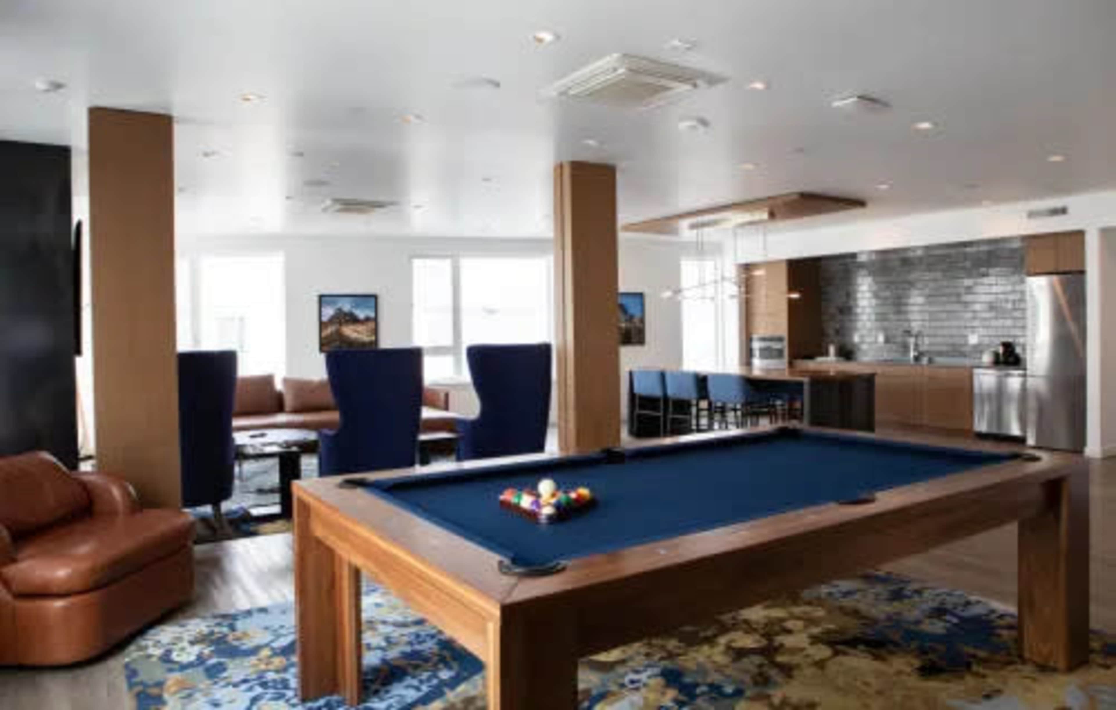 A modern interior lounge area featuring a pool table, a seating area with large chairs, and a kitchen in the background.
