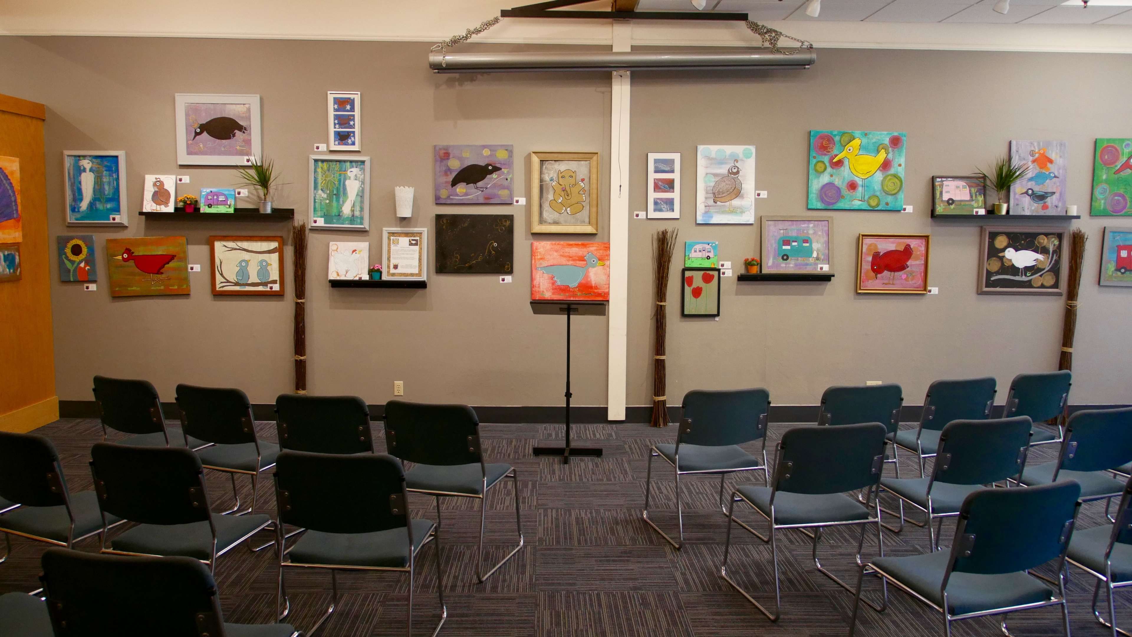 A gallery space with several colorful framed artworks displayed on the walls and empty chairs arranged in rows facing a stand.