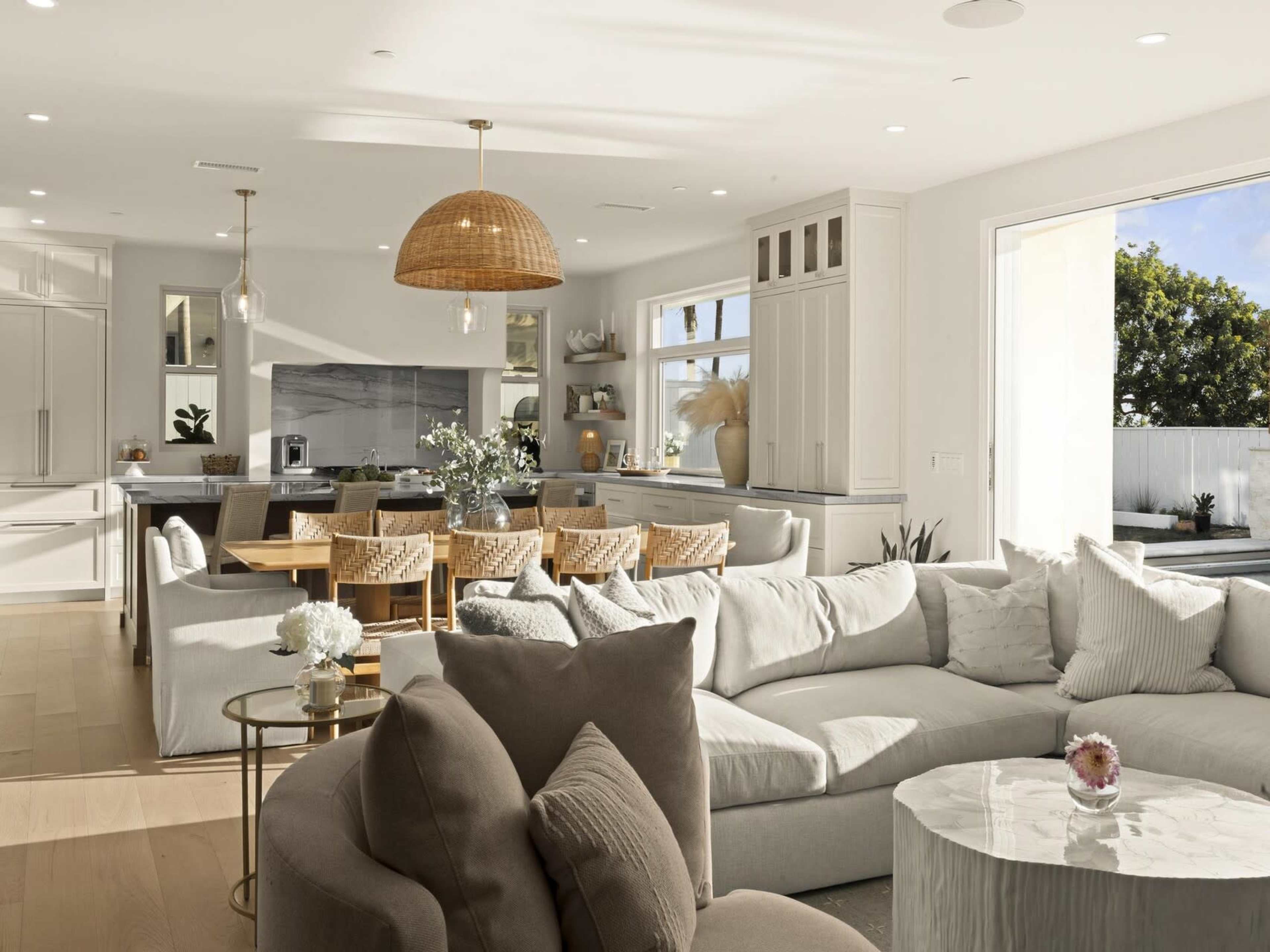 The image shows a modern, open-concept living space featuring a large sectional sofa, a glass coffee table, and a dining area with woven chairs and a wooden table.