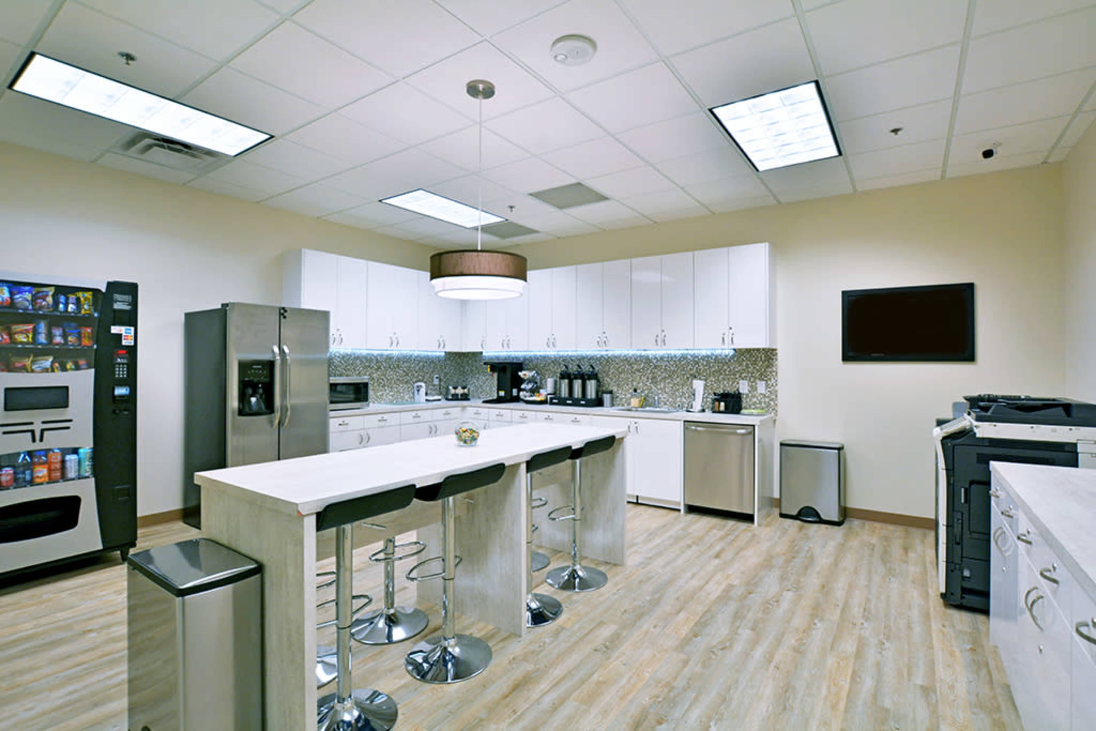 The image shows a modern kitchen area with a large island, stainless steel appliances, a vending machine, and a copier station.
