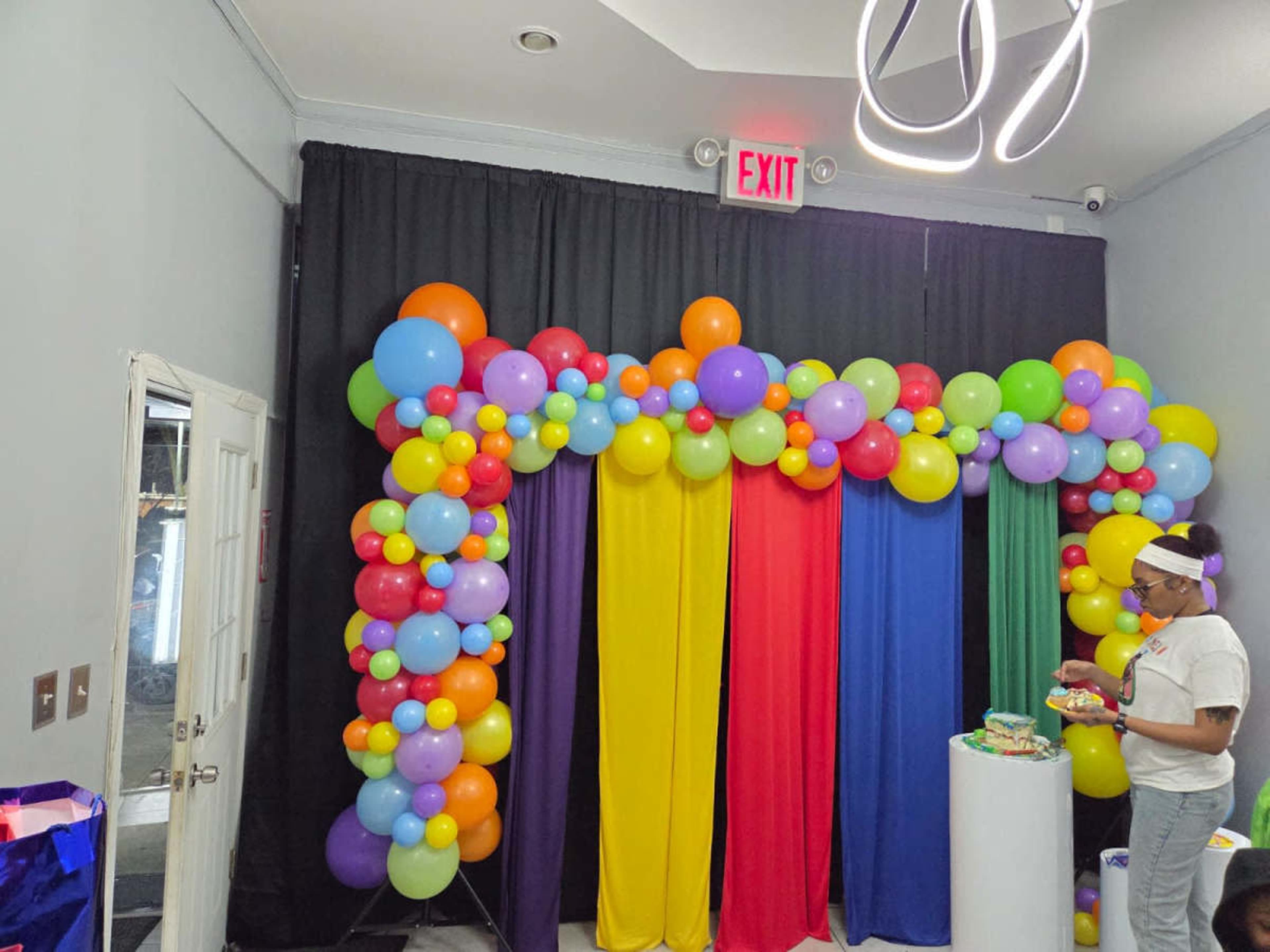 The image shows a colorful balloon arch with red, yellow, green, purple, and blue balloons above a backdrop of draped fabric in the same colors, set against a dark curtain.