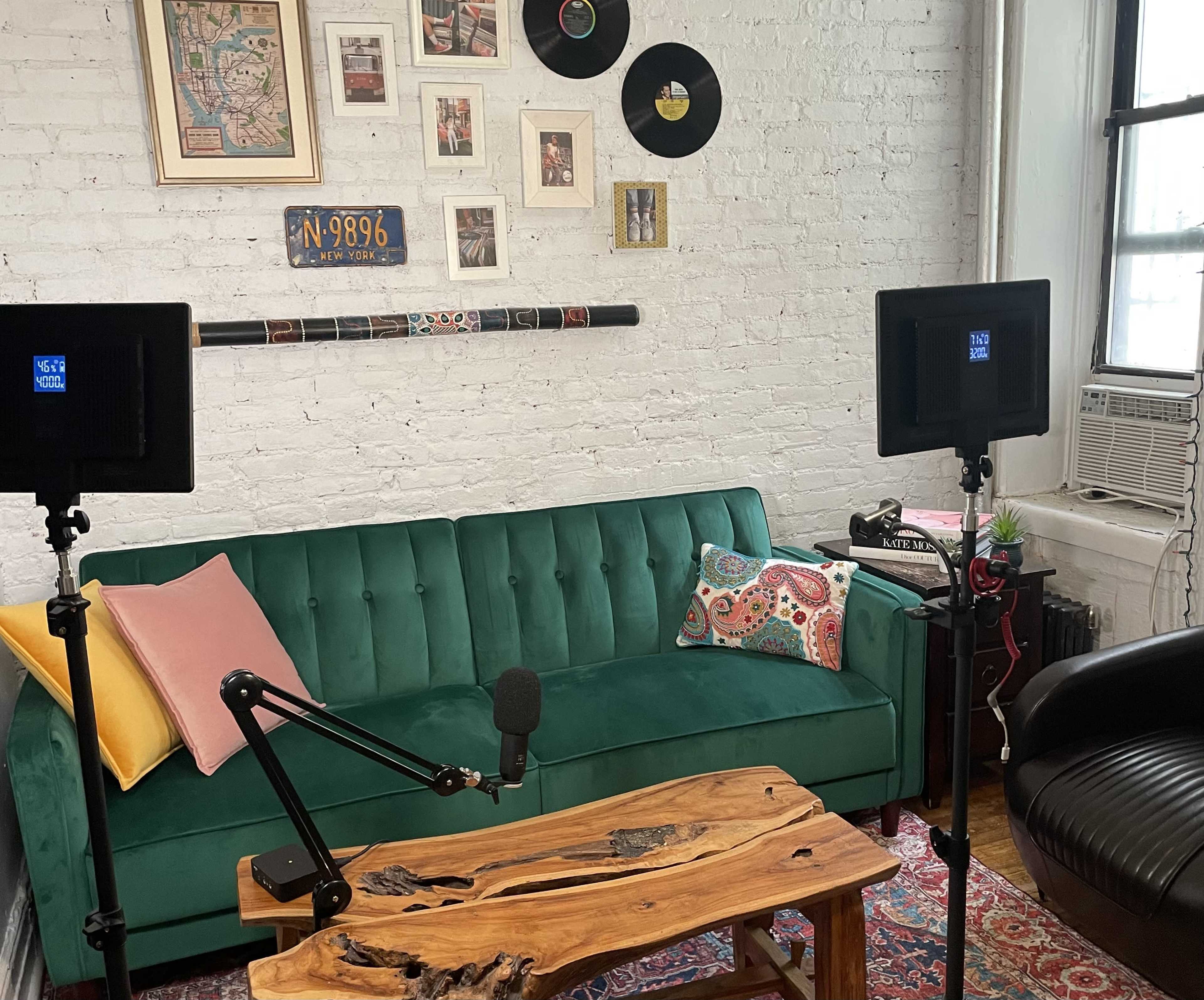 A stylish living room features a green velvet couch, artwork on the walls, a wooden coffee table, and two microphone stands with lights positioned for recording.
