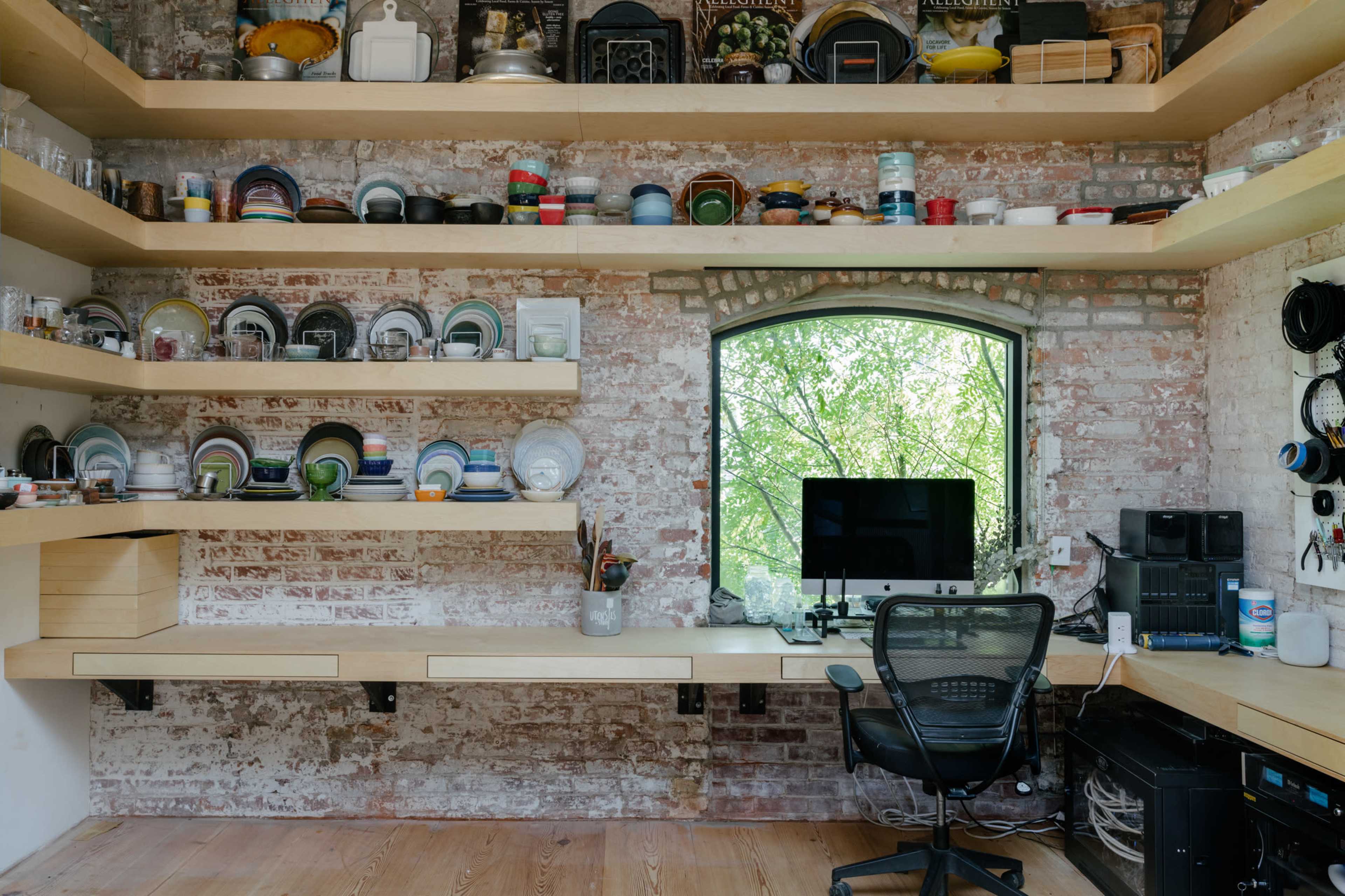 A workspace features a desk in front of a large window, surrounded by shelves filled with various dishes and kitchen items, against a brick wall.