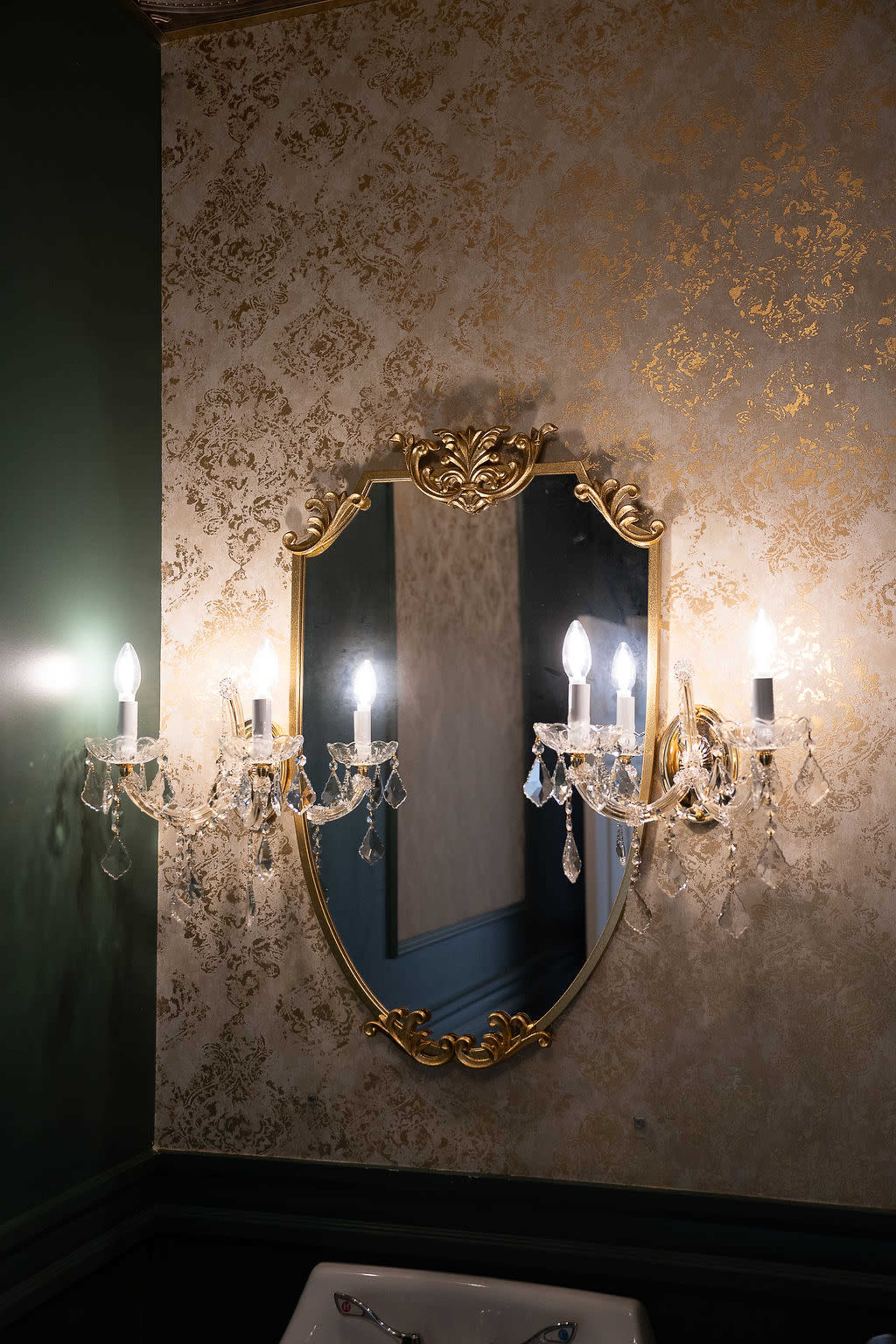 The image shows a decorated bathroom with a large ornate mirror flanked by two crystal wall sconces illuminated by light bulbs.