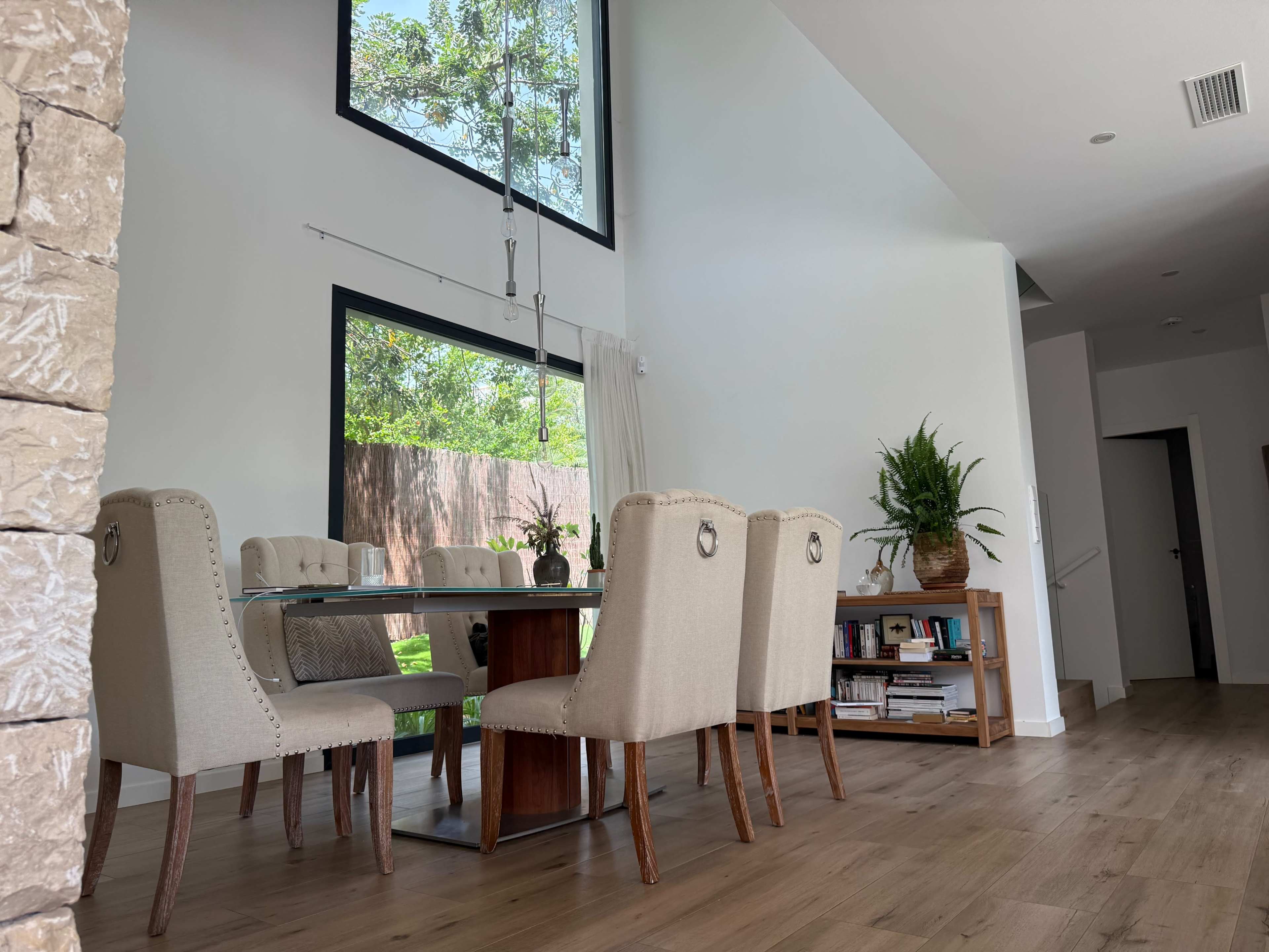 A modern dining area features a wooden table surrounded by upholstered chairs, large windows letting in natural light, and a plant-filled corner shelf.