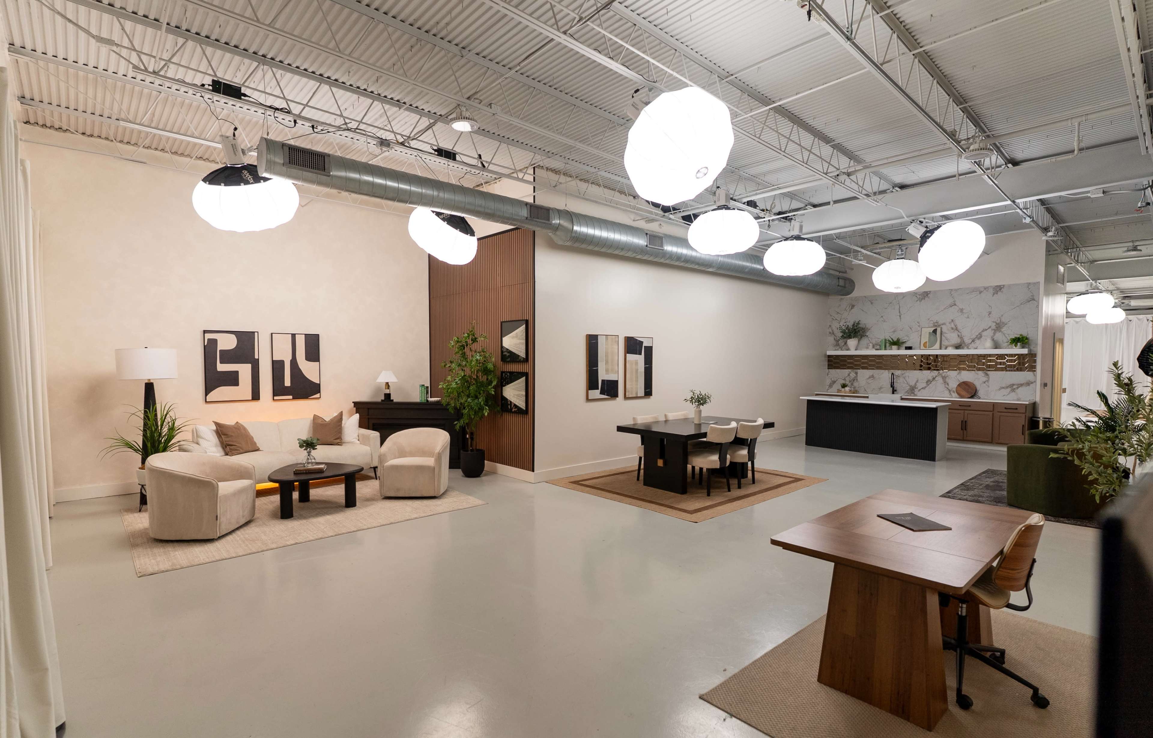 The image shows a spacious, modern interior featuring distinct seating areas, artwork on the walls, and various plants, along with a kitchen in the background.