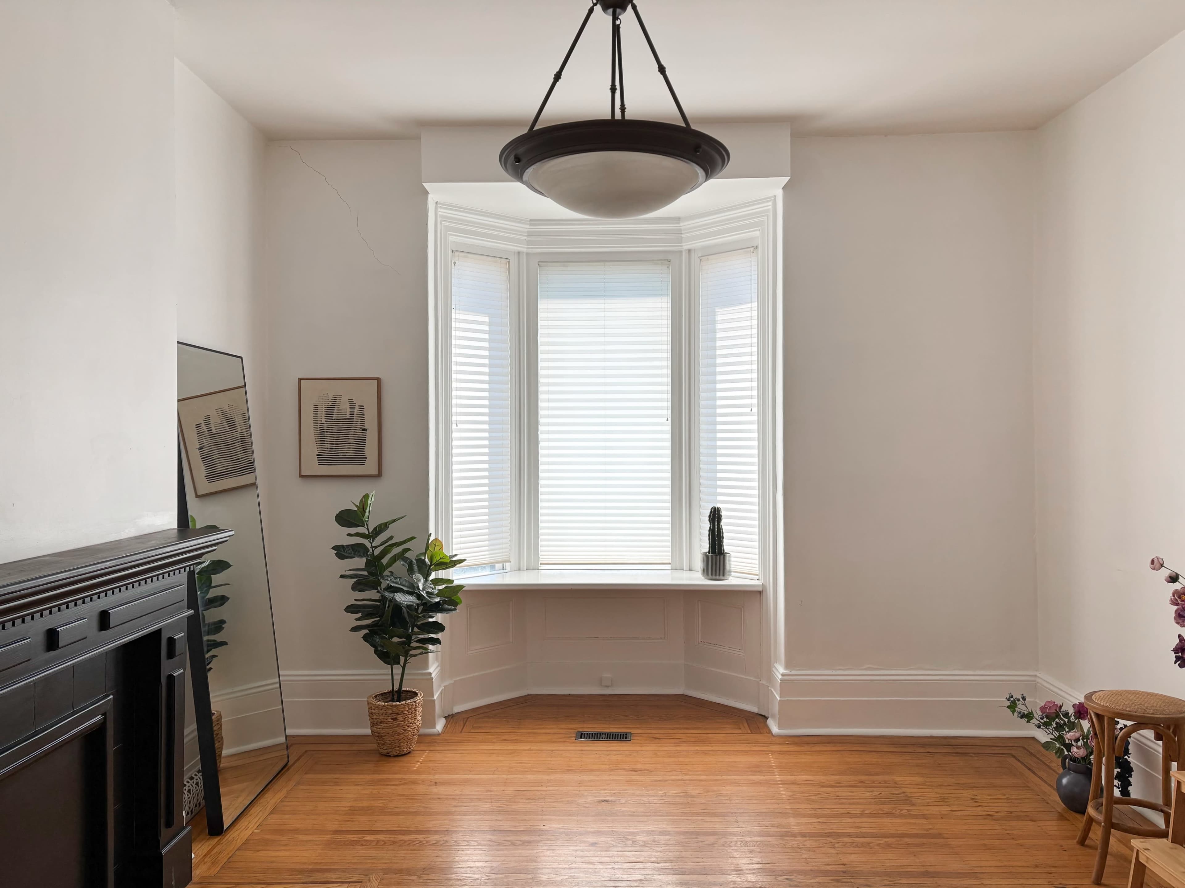 The image shows a bright, empty room with a large bay window, a potted plant in the corner, and a mirrored wall art.