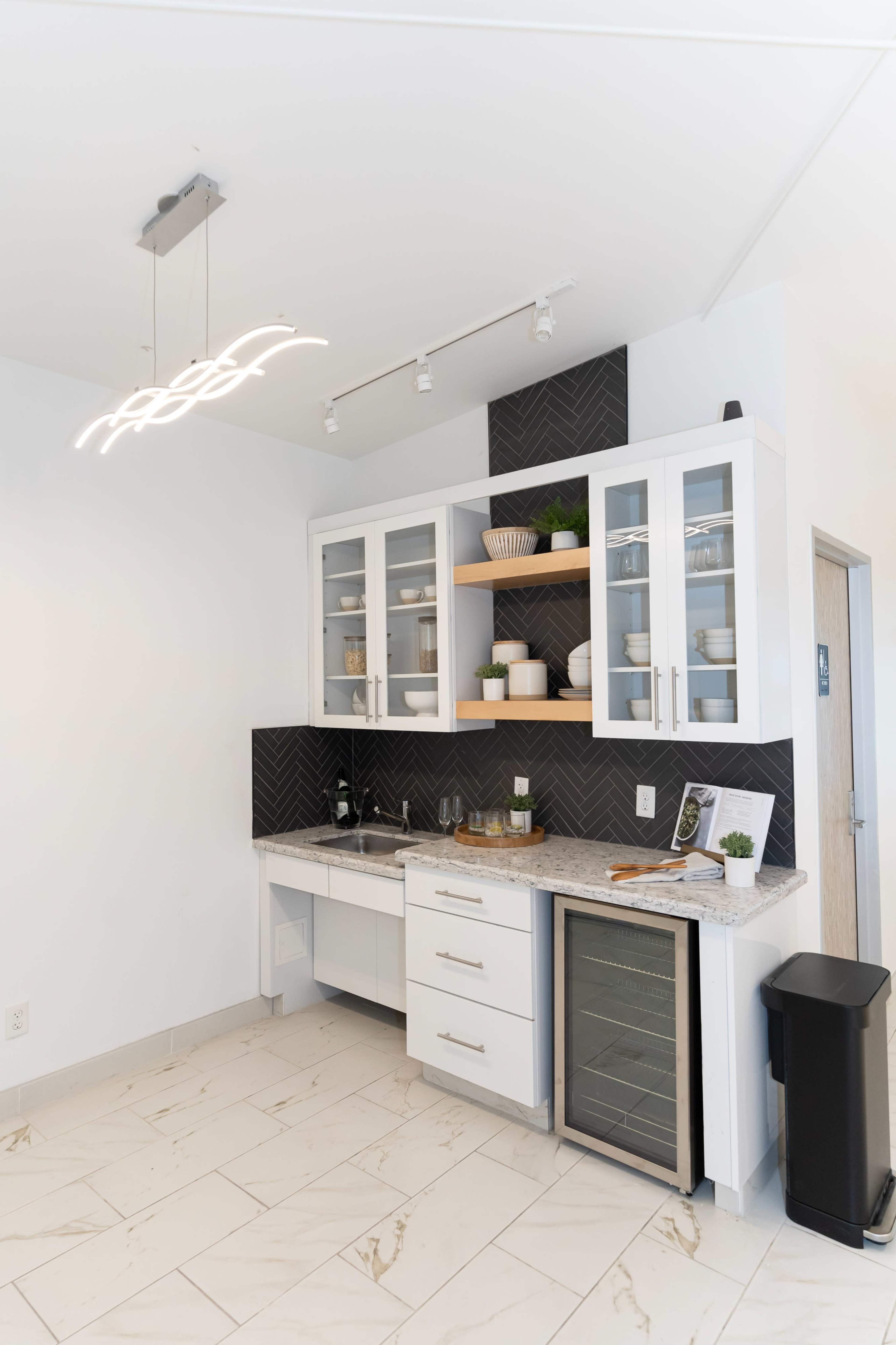 The image shows a modern kitchen corner with white cabinetry, a granite countertop, a small sink, a wine fridge, and a decorative backsplash.
