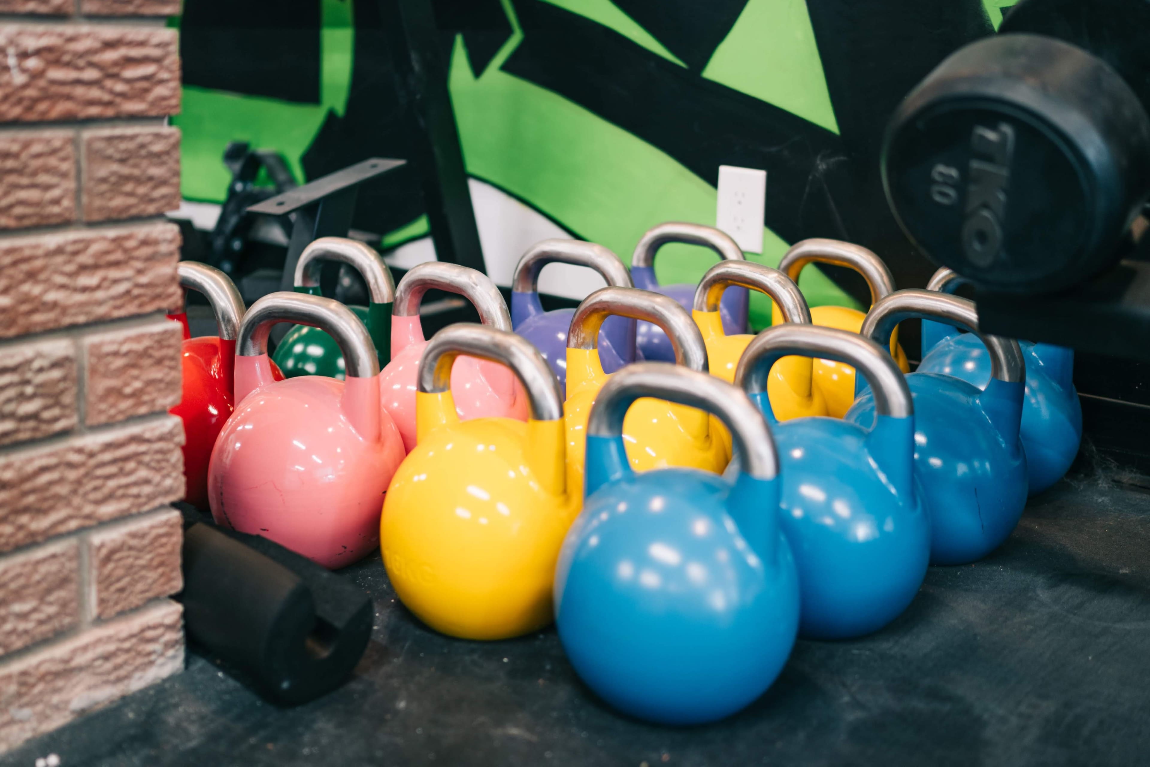 A row of colorful kettlebells is arranged on a gym floor beside a wall decorated with green and black designs.