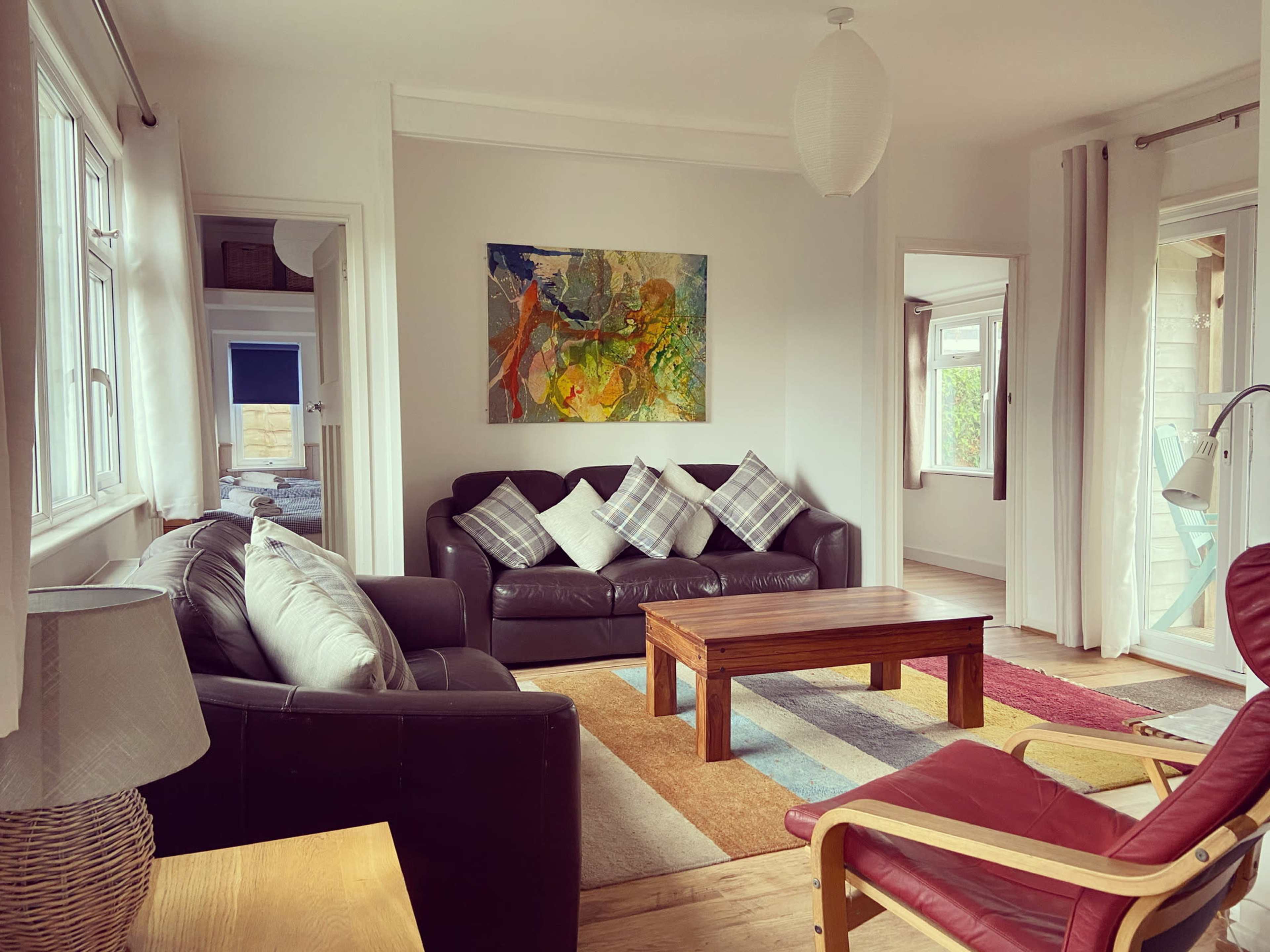 The room features a black leather sofa with plaid throw pillows, a wooden coffee table, and an abstract painting on the wall, with large windows letting in natural light.