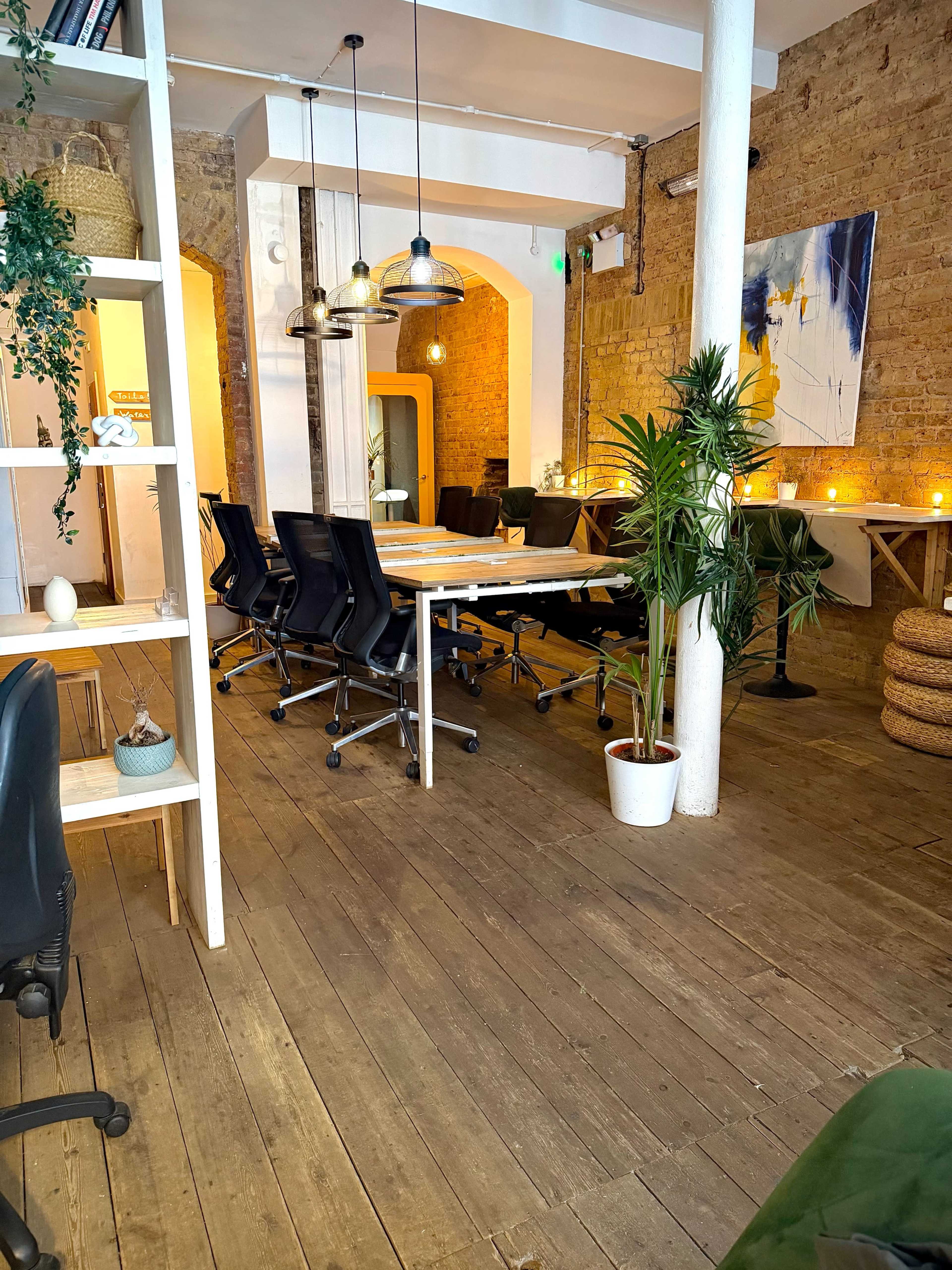 The image shows a modern, open-concept office space featuring a large table, black ergonomic chairs, and decorative plants, with warm lighting enhancing the brick walls.
