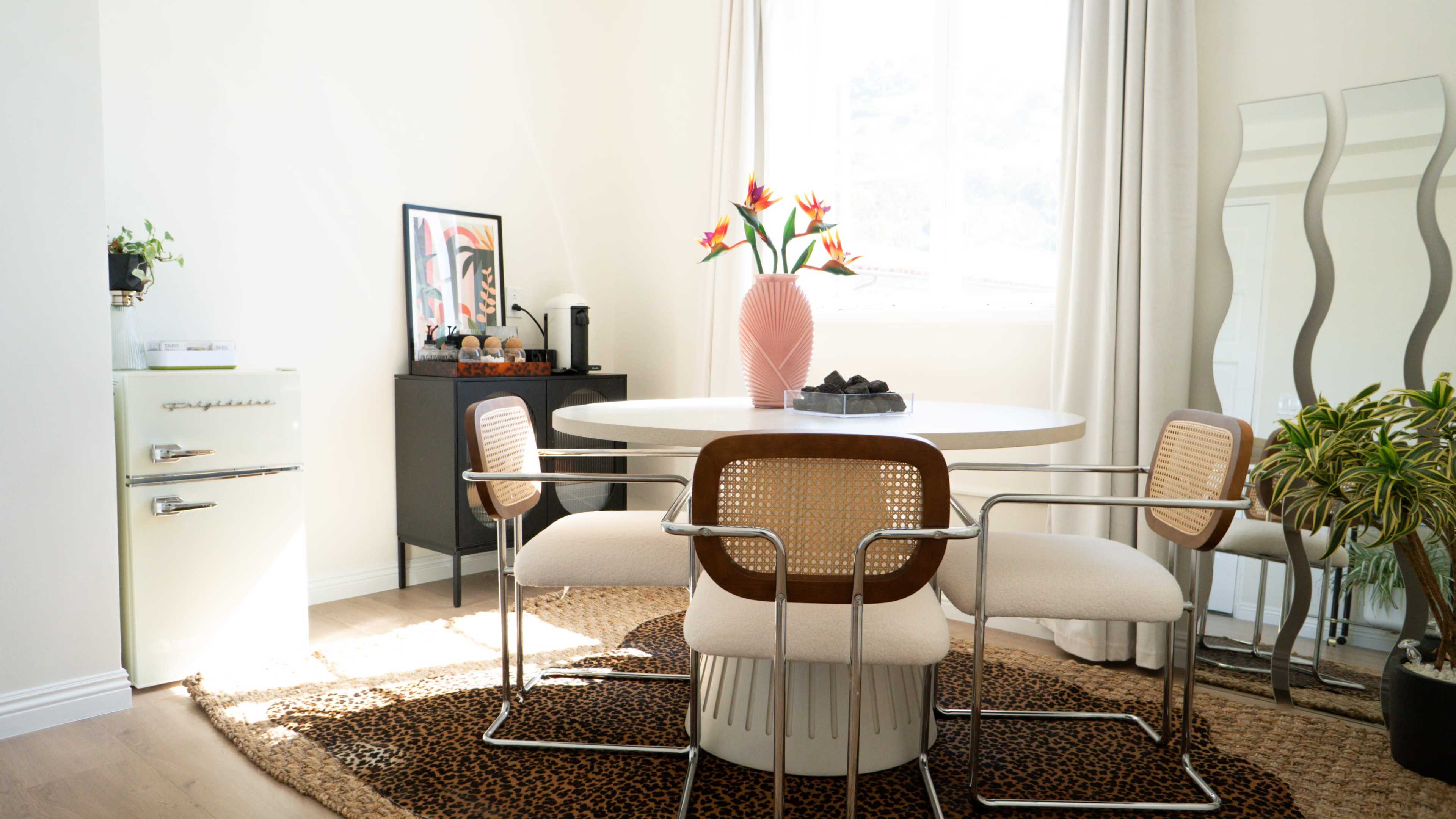 A round dining table surrounded by four chairs sits in a bright room, adorned with a vase of flowers and framed artwork, next to a vintage refrigerator and a textured rug.